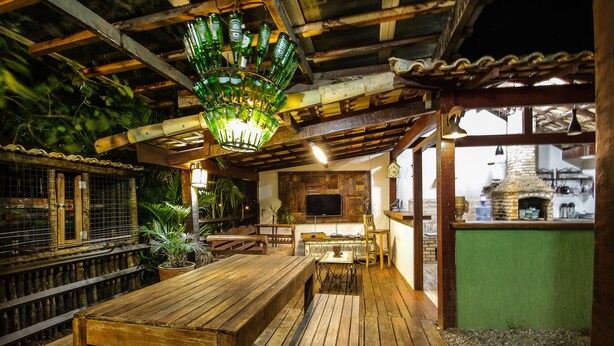A rustic wooden dining area with a bench and table, a hanging light fixture made of green glass bottles, and a brick oven in the background. - Pousada NOVA ESTRELA INN (Búzios)
