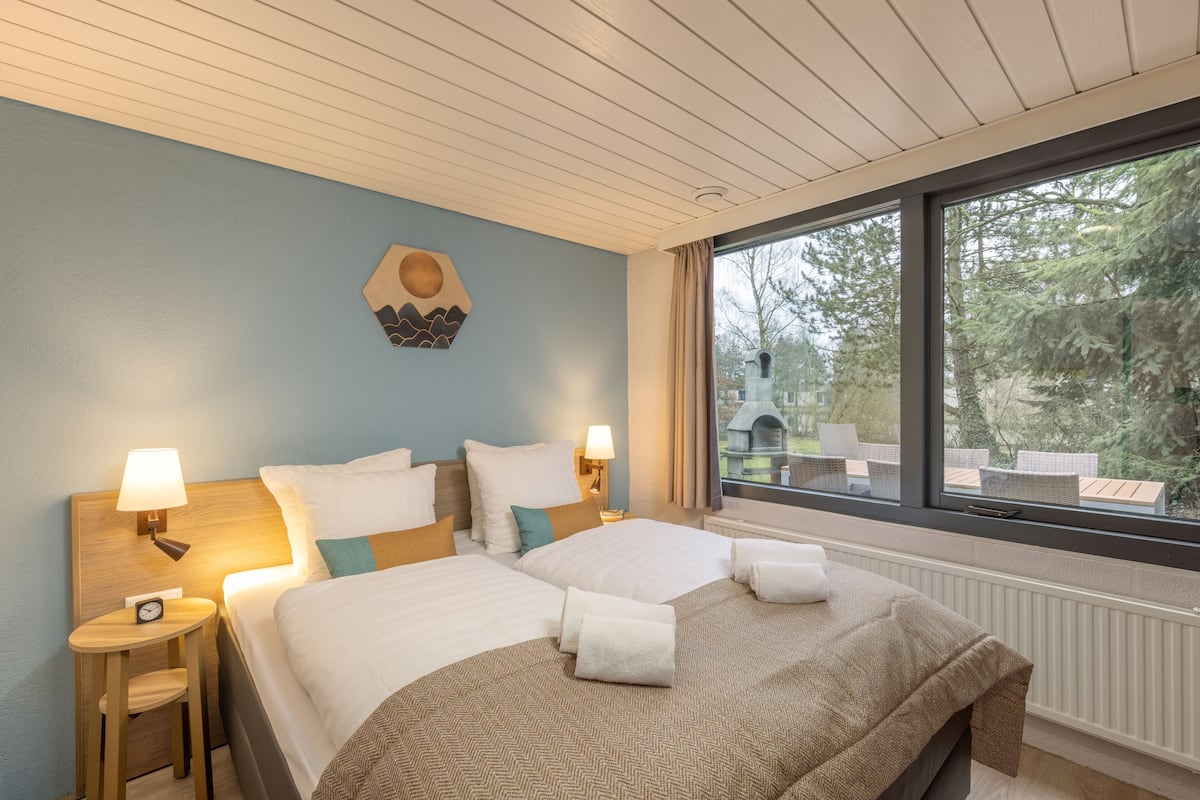 Habitación de hotel con dos camas, cabecero de madera, lámparas de noche, un reloj y ventana con vistas a árboles y mobiliario exterior.