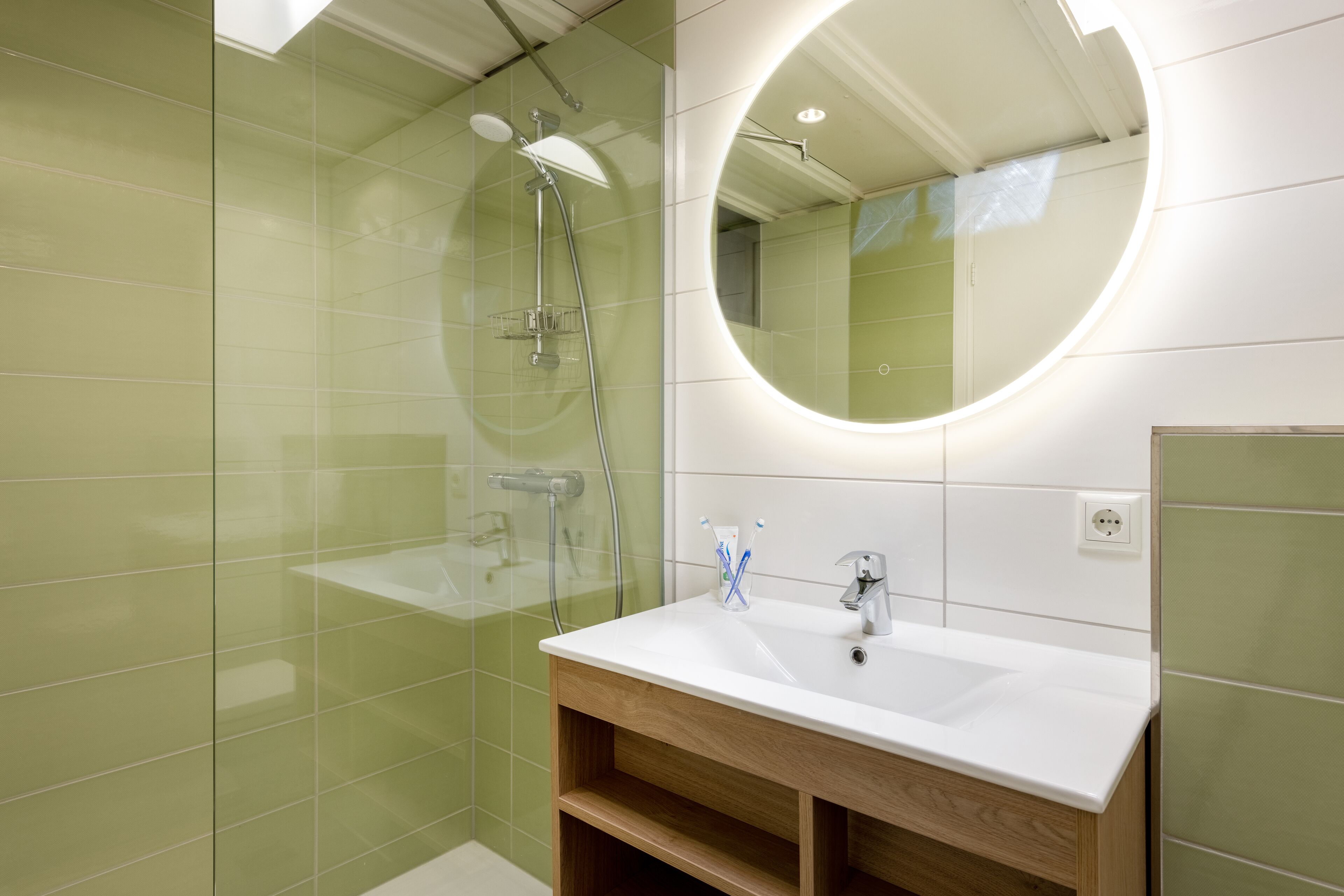 A modern bathroom with a glass-enclosed shower, a white sink with a wooden vanity, and a round mirror with a light.