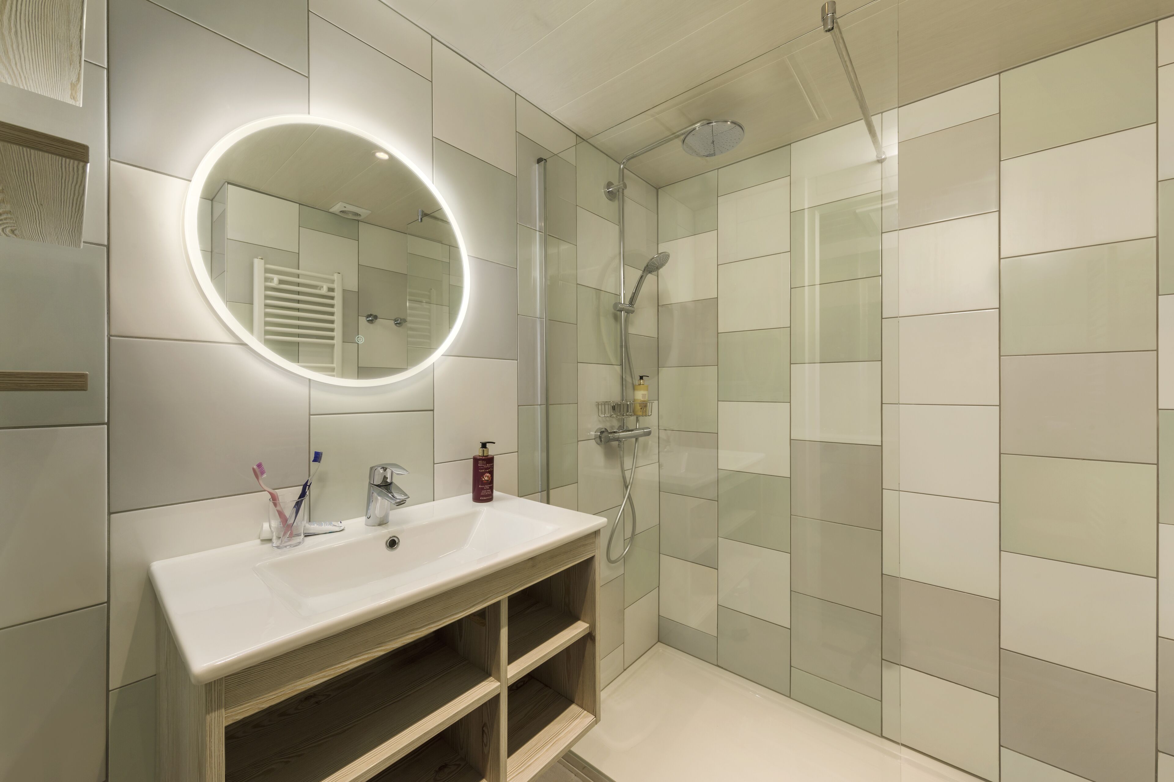 A modern bathroom with a white sink, a mirror with a circular light, and a glass-enclosed shower.