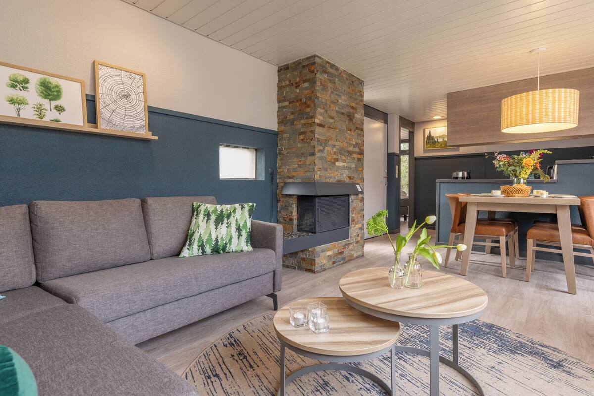 Una sala de estar moderna con un sofá gris, una chimenea de piedra, una zona de comedor con una mesa y sillas de madera, y una lámpara colgante.