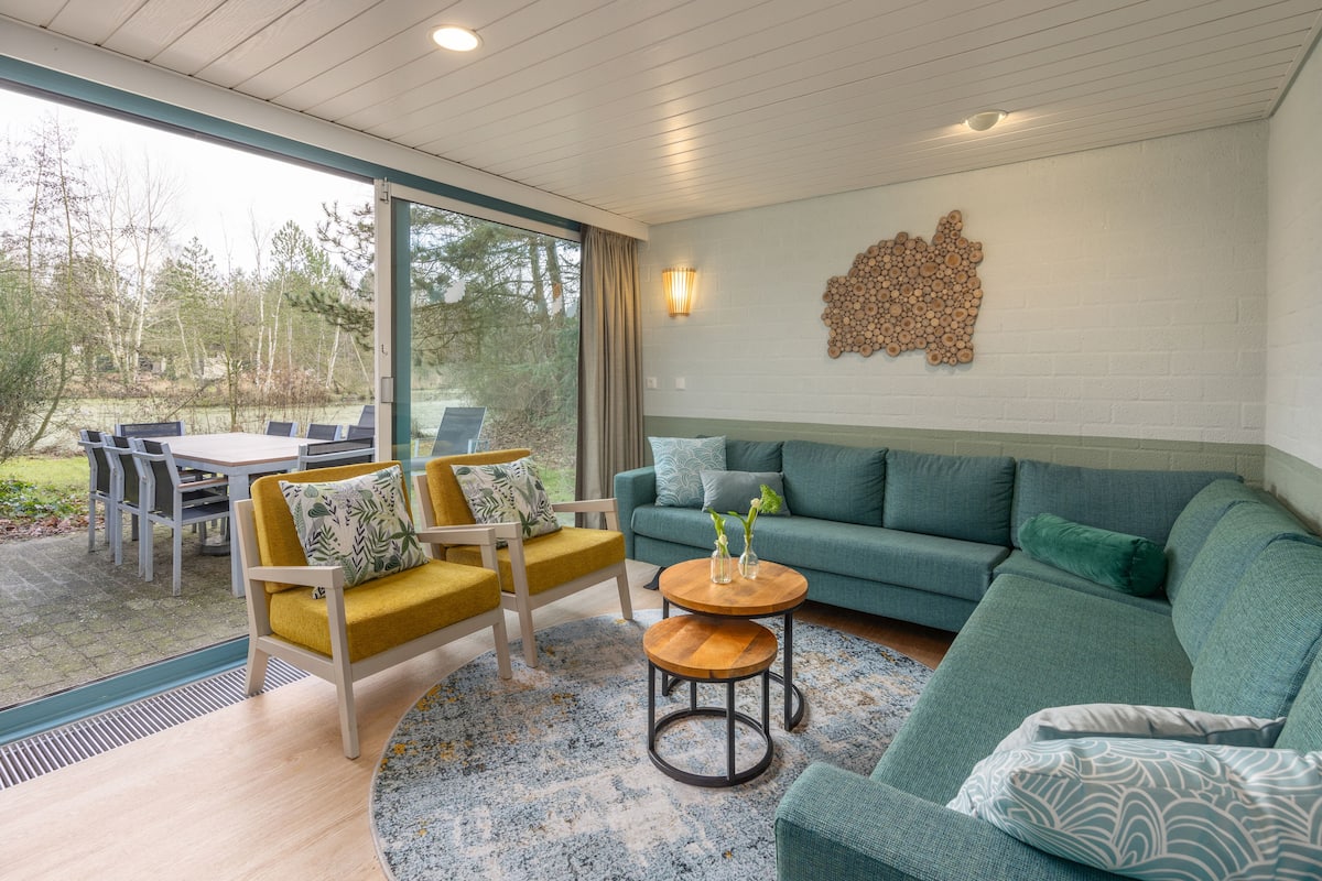 Una sala moderna con un sofá verde, sillones amarillos, una mesa de centro de madera y una zona de comedor con mesa y sillas.