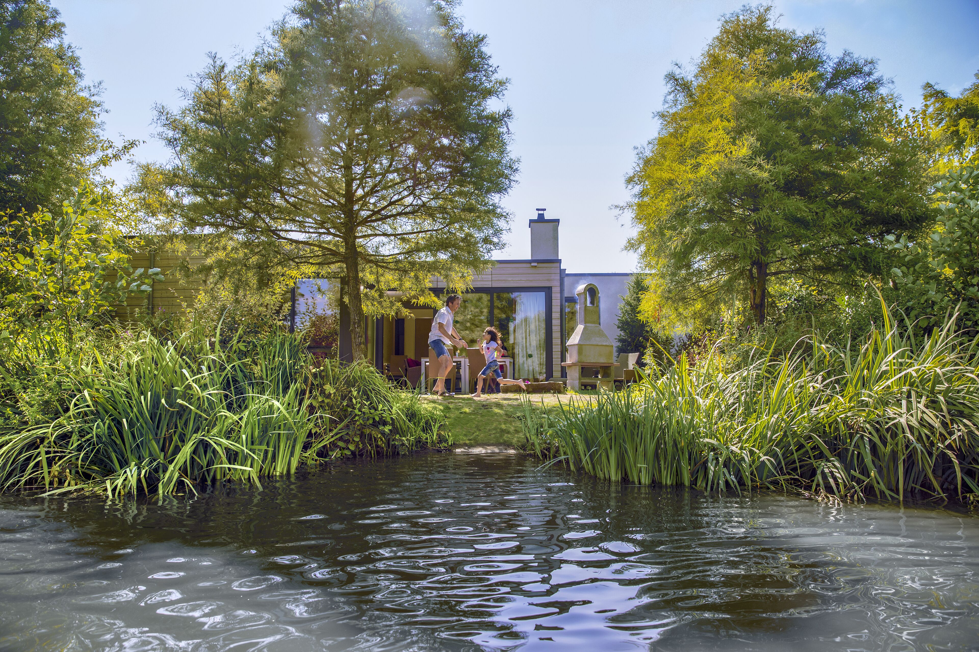 A house with a water feature in front, surrounded by tall grass and trees.