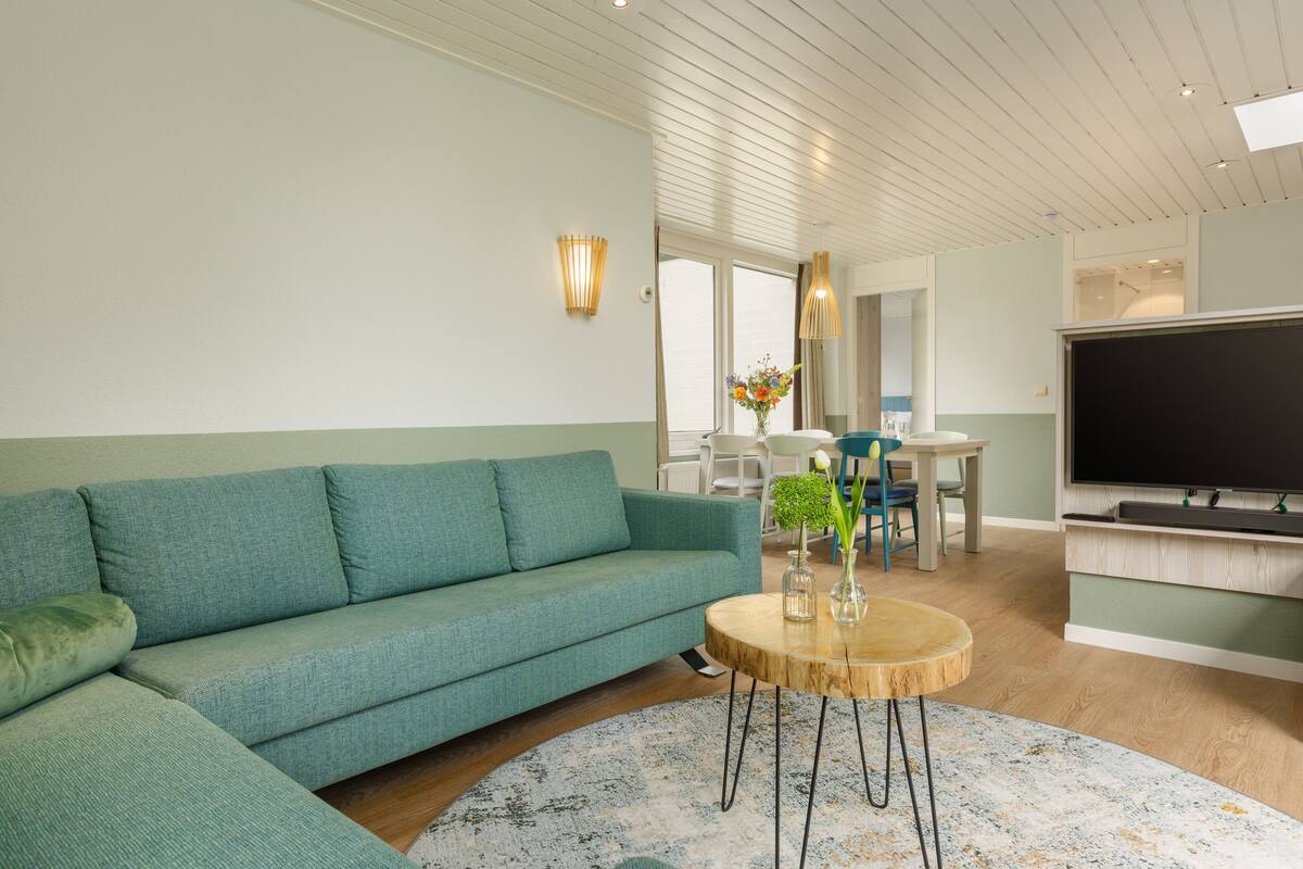 A modern living room with a teal sofa, a round wooden coffee table, and a flat-screen TV.
