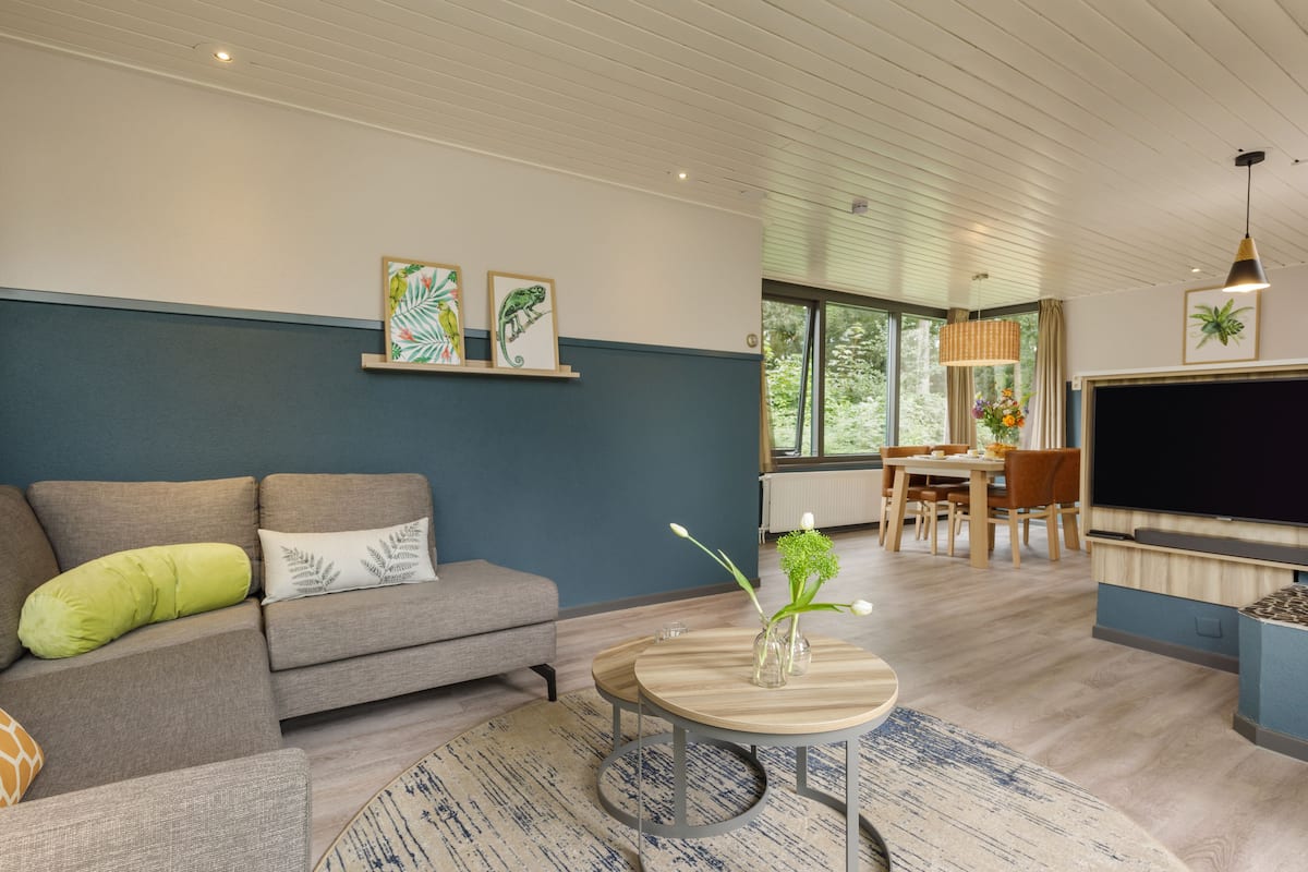 A modern living room with a grey sofa, a round coffee table, and a flat-screen TV. There is a dining area with a table and chairs, and a large window providing natural light.