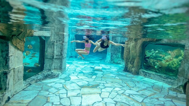 An underwater view of a stone-tiled pool with two people swimming, surrounded by fish. - Center Parcs Het Heijderbos (Heijen)