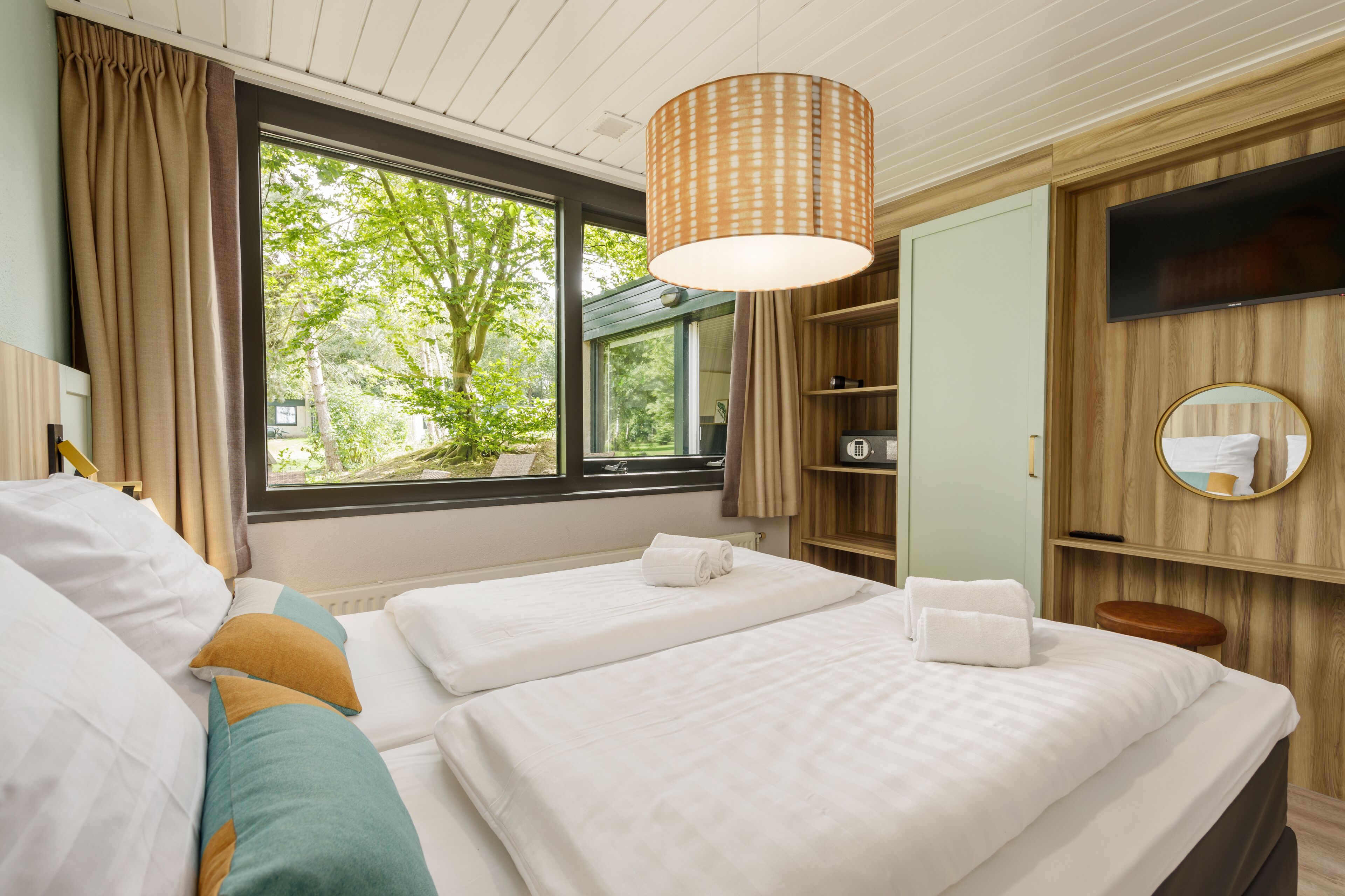 A hotel room with two beds, a large window with a view of greenery, a wooden wardrobe, and a television mounted on the wall.
