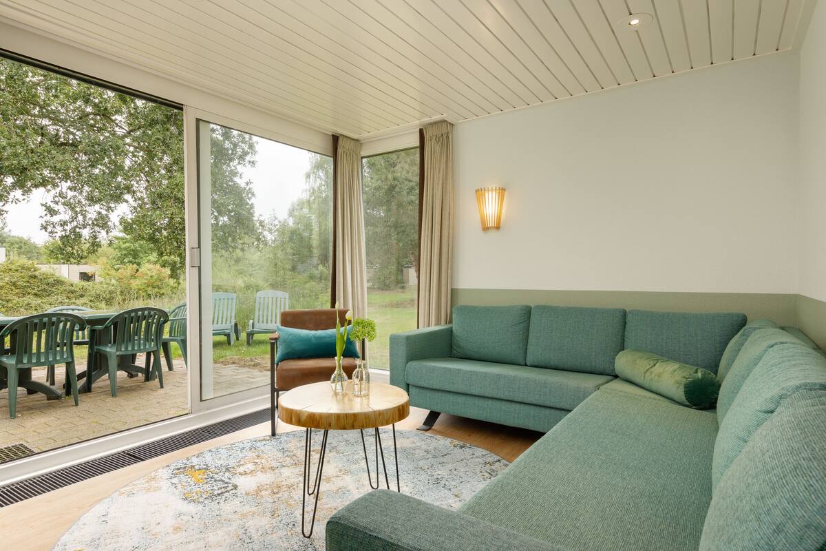 A modern living room with a green sofa, a round wooden coffee table, and a view of the outdoors through large sliding glass doors.