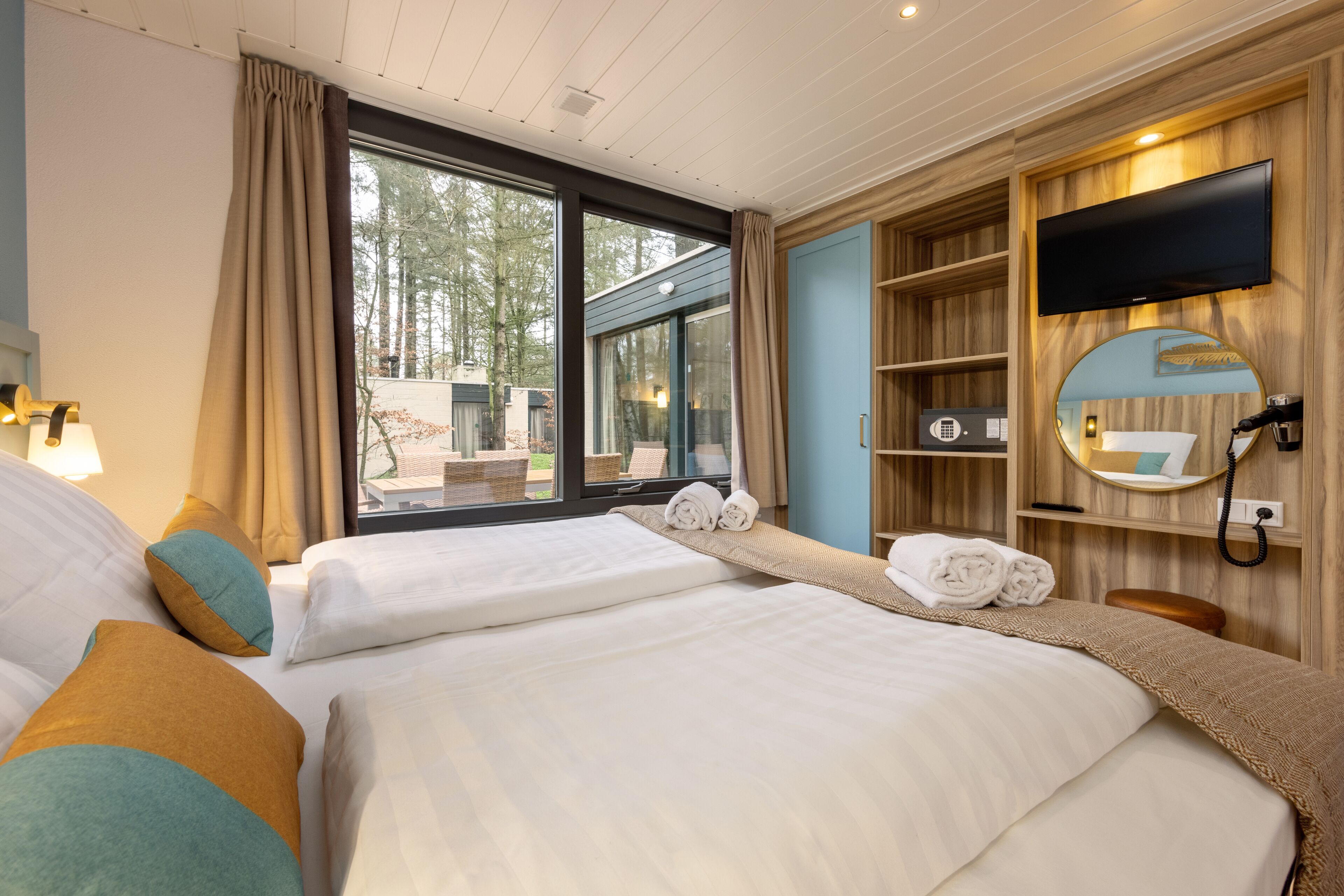 A hotel room with two beds, a TV, a mirror, and a window with a view of trees.