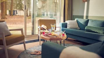 A modern living room with a blue sofa, a wooden coffee table with snacks and drinks, a white armchair, and a large window with a view of trees.