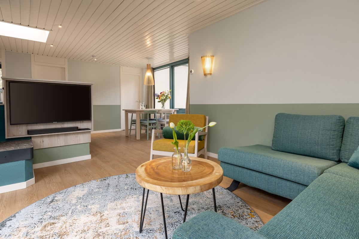 A modern living room with a flat-screen TV, a round wooden coffee table, teal sofas, and a dining area with a table and chairs.