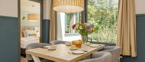 A dining area with a wooden table set for four, a vase with flowers, and a view of greenery outside.
