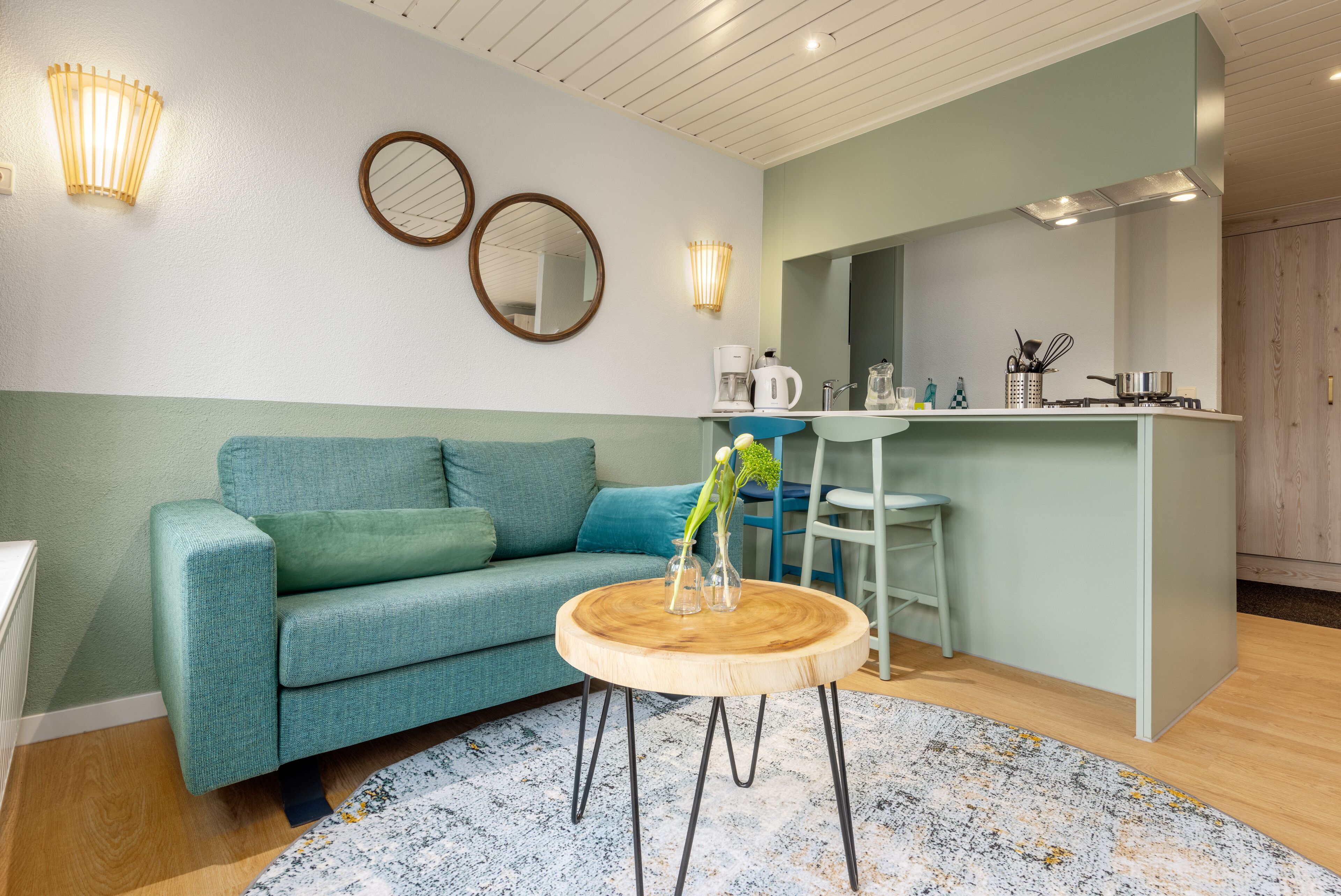 Ein modernes Wohnzimmer mit einem türkisfarbenen Sofa, einem Holztisch und einer Küchenzeile mit Spüle und Herd.