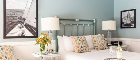 A bedroom with a bed, two bedside lamps, a framed picture of a sailboat, a vase with flowers, and a bottle of wine on a tray.