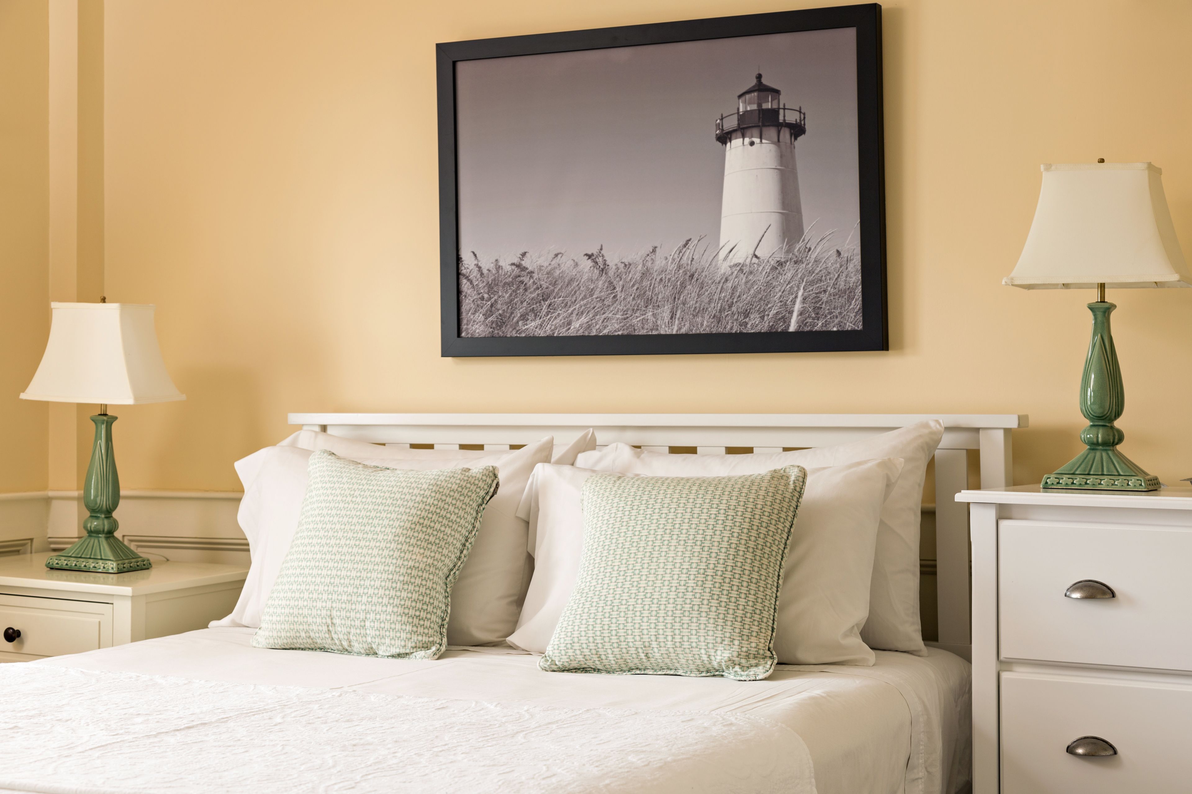 A neatly made bed with white linens and two pillows, a framed picture of a lighthouse on the wall, and two bedside tables with lamps.