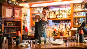 A bar with a bartender pouring a drink into a glass. There are various bottles and glasses on shelves in the background.