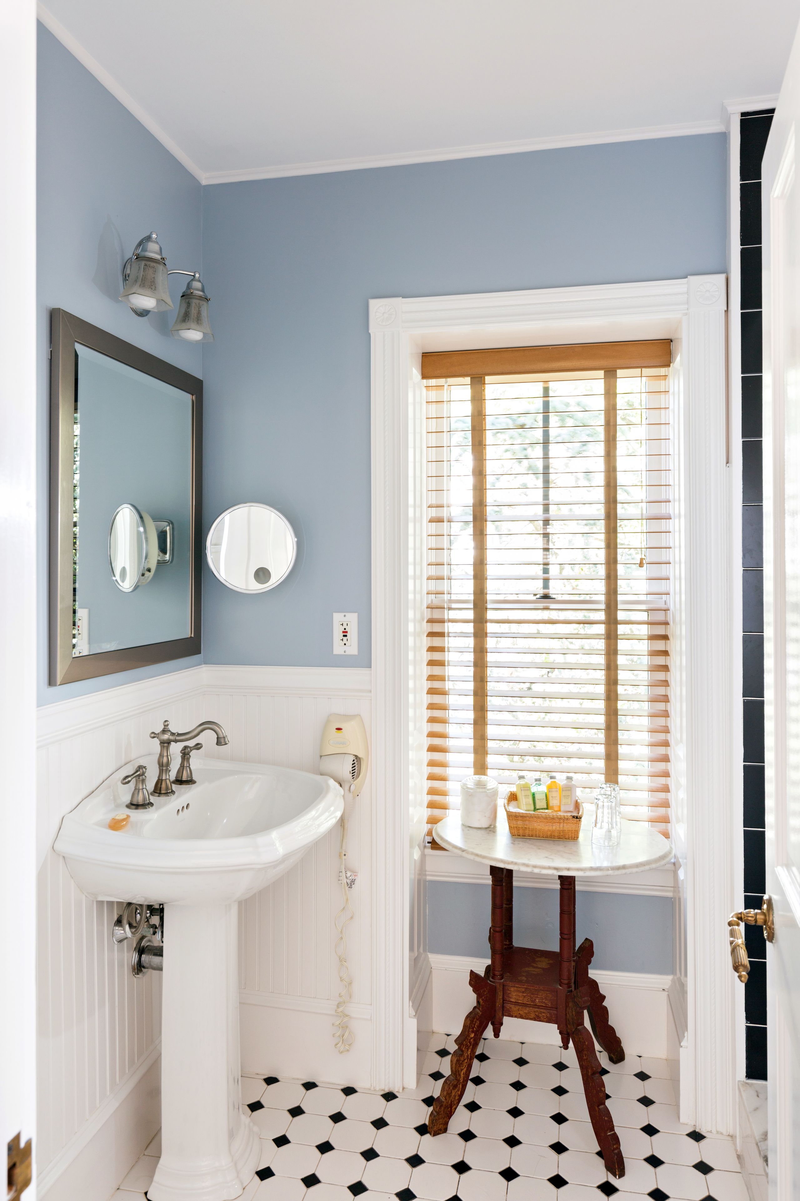 Un baño con lavamanos blanco, espejo y ventana con persianas.