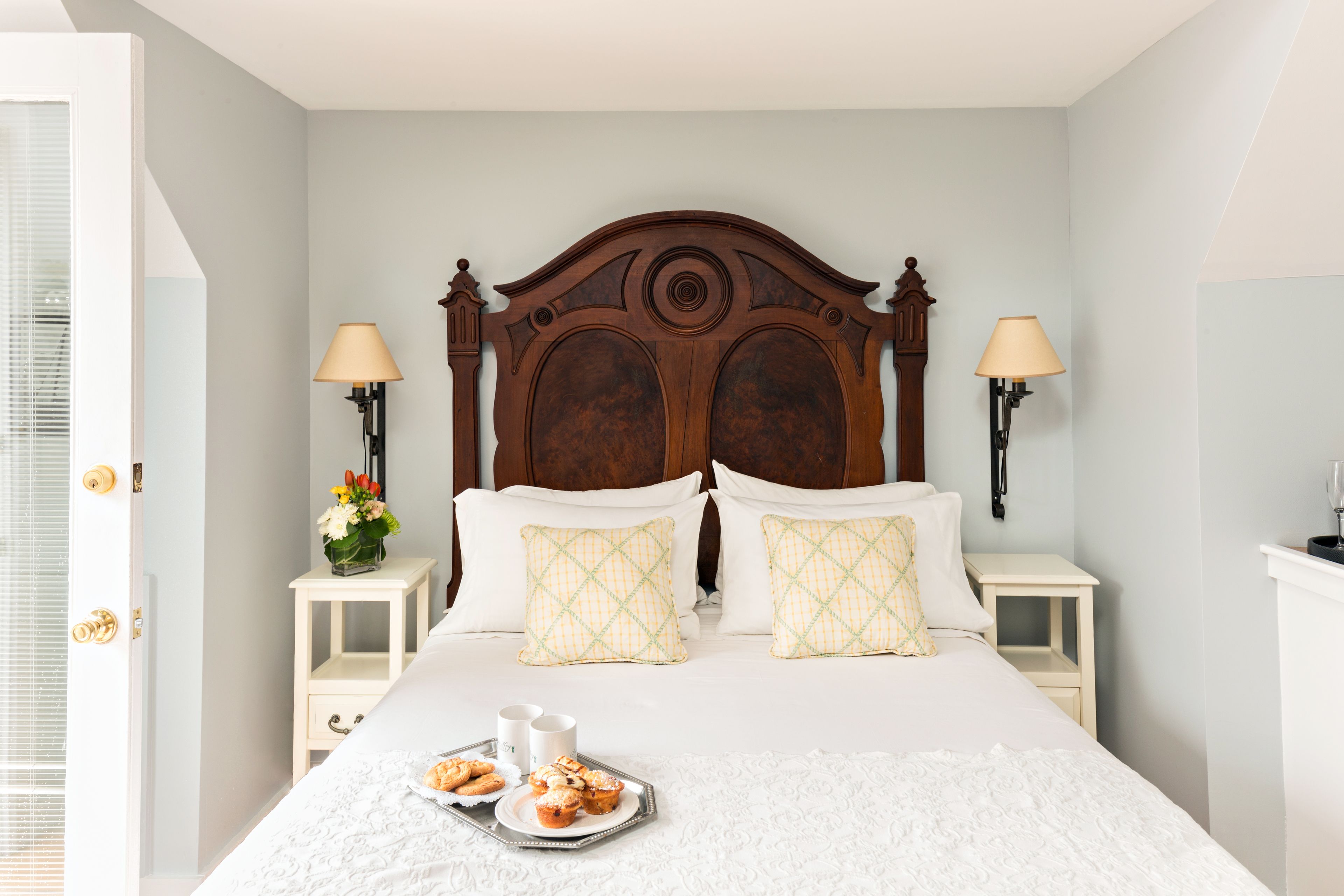 A neatly made bed with a wooden headboard, two bedside tables with lamps, and a tray of pastries on the bed.