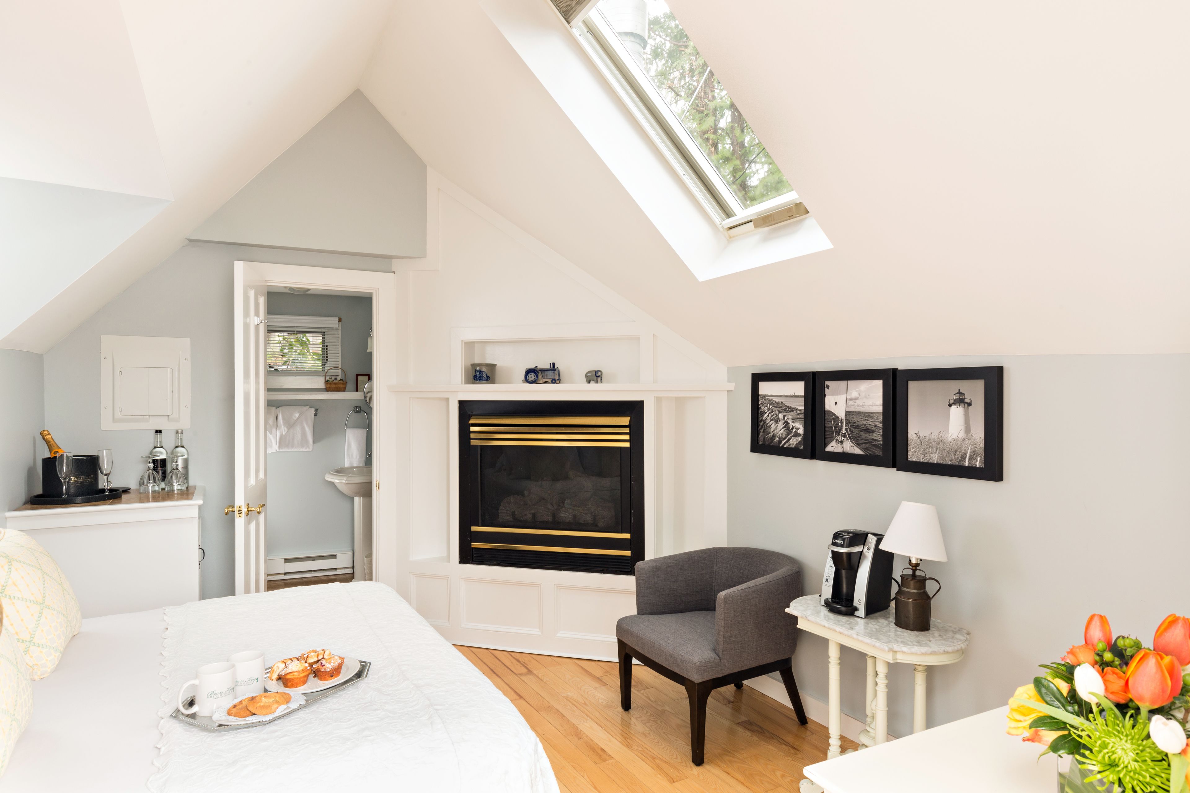 A bedroom with a skylight, a fireplace, a bed with a tray of pastries, a chair, and a small table with a lamp and a vase of flowers.