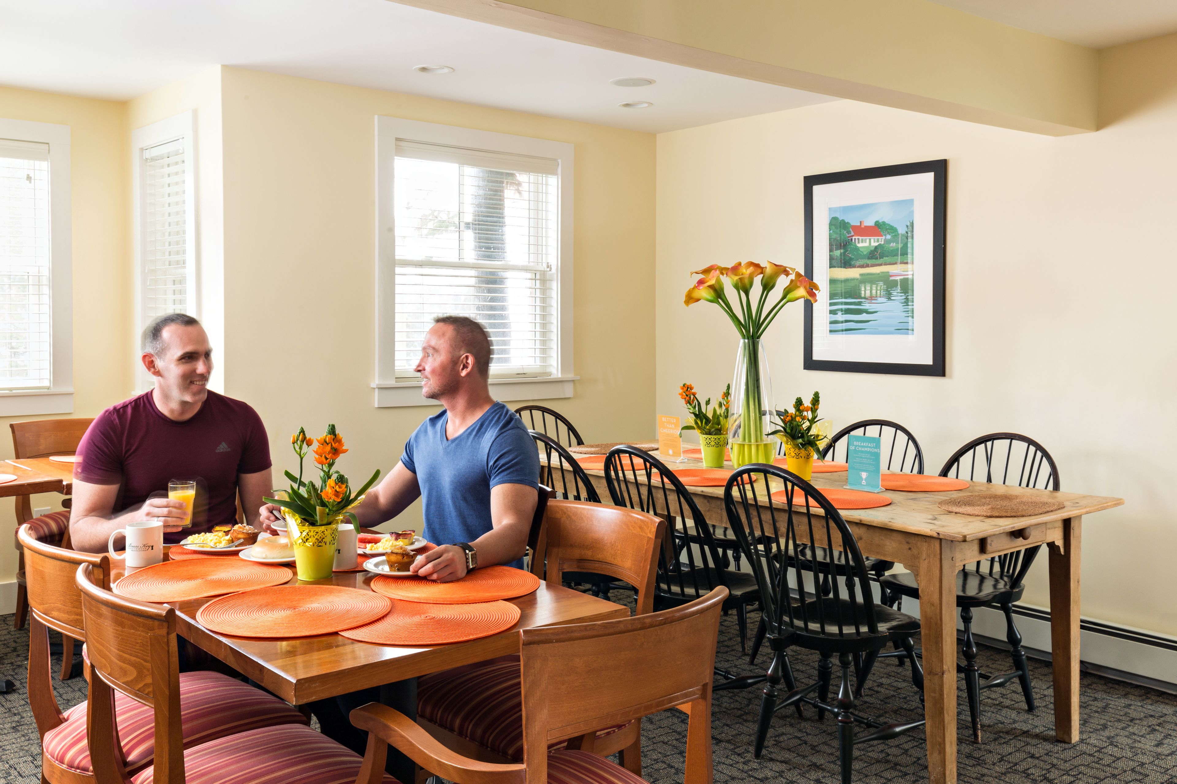 A dining area with wooden tables and chairs, a vase with flowers, and a framed picture on the wall.