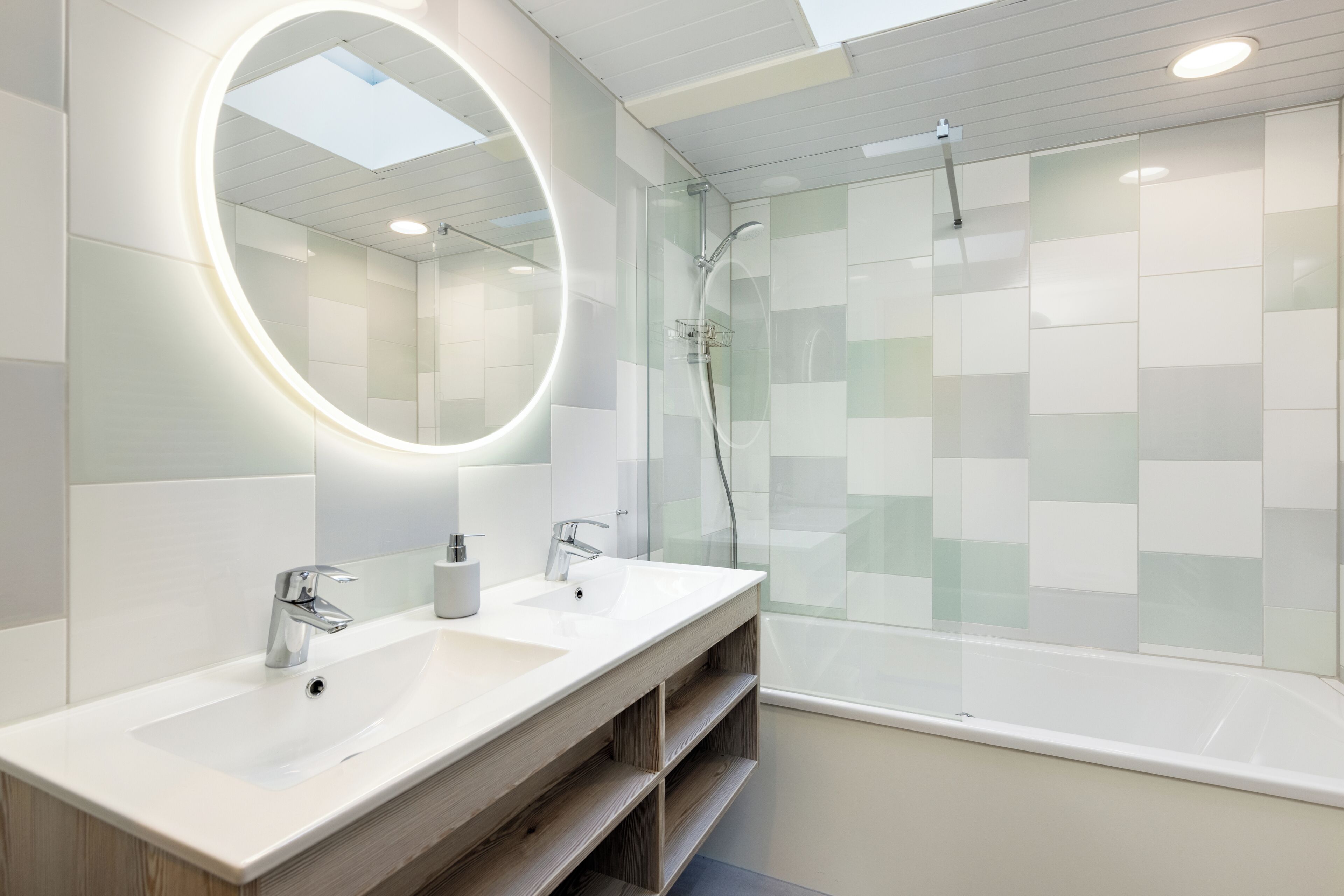 A modern bathroom with a large oval mirror, two sinks, a glass-enclosed shower, and a skylight.