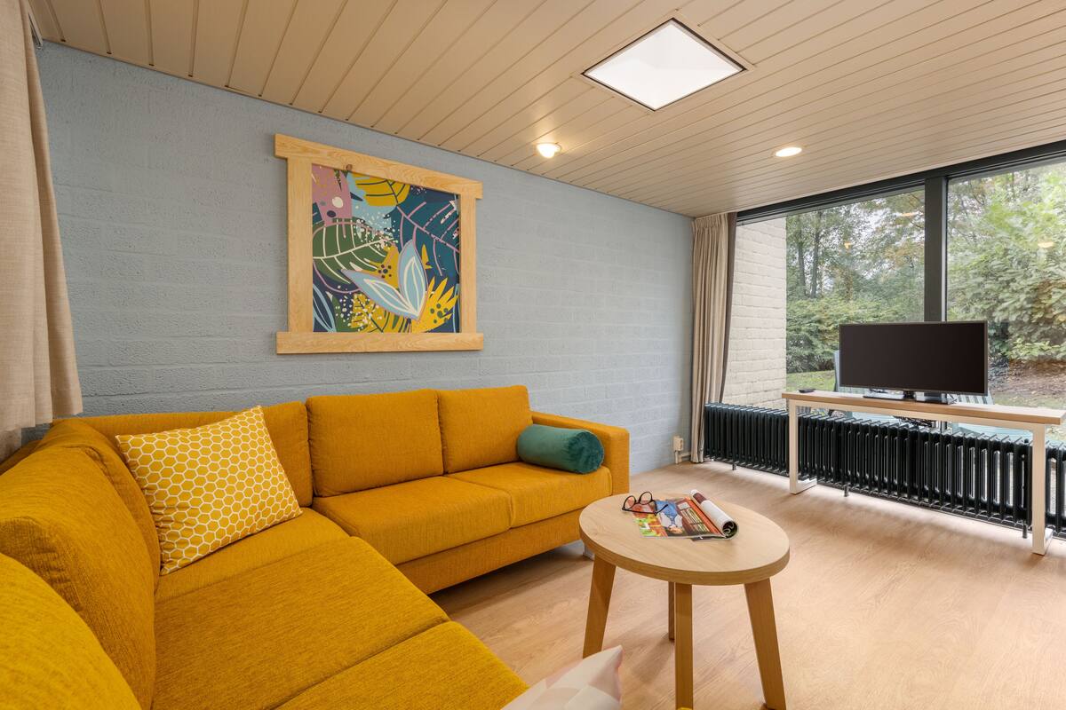 A modern living room with a mustard sofa, a round wooden coffee table, and a large window offering a view of trees.