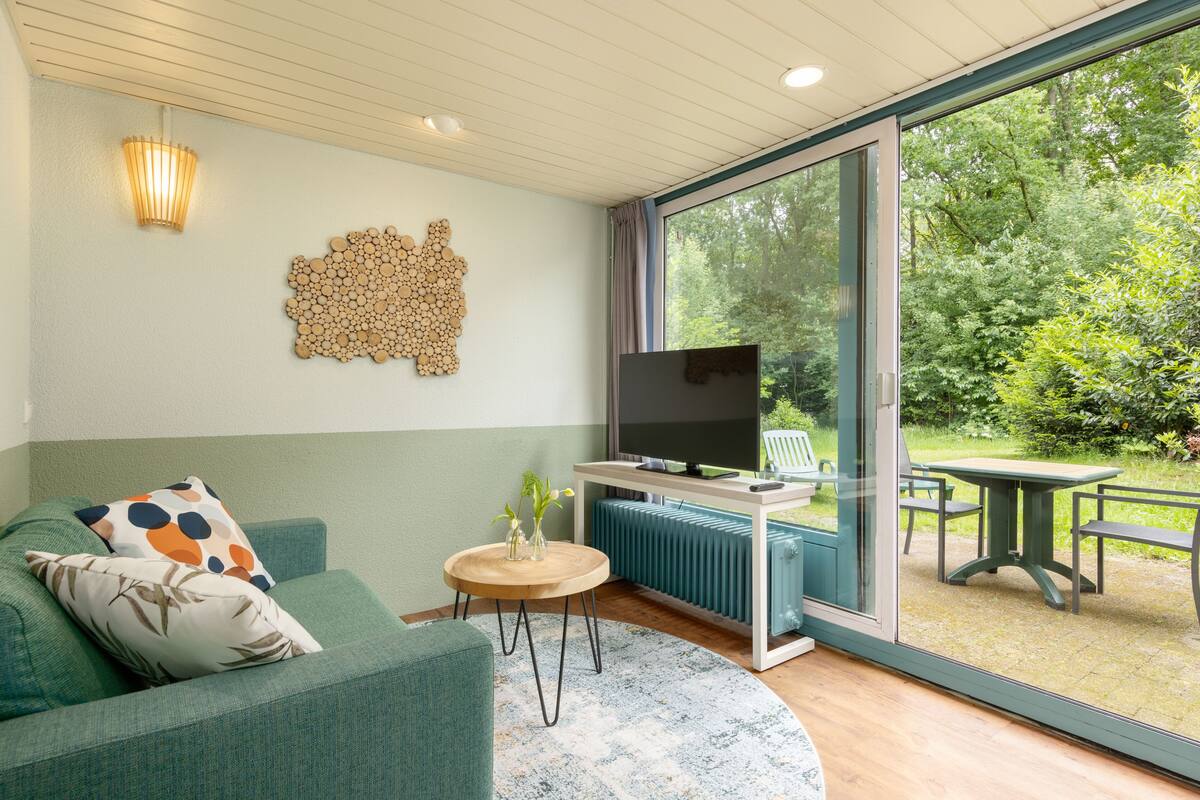 A modern living room with a sofa, a coffee table, a TV, and a view of a garden through large windows.