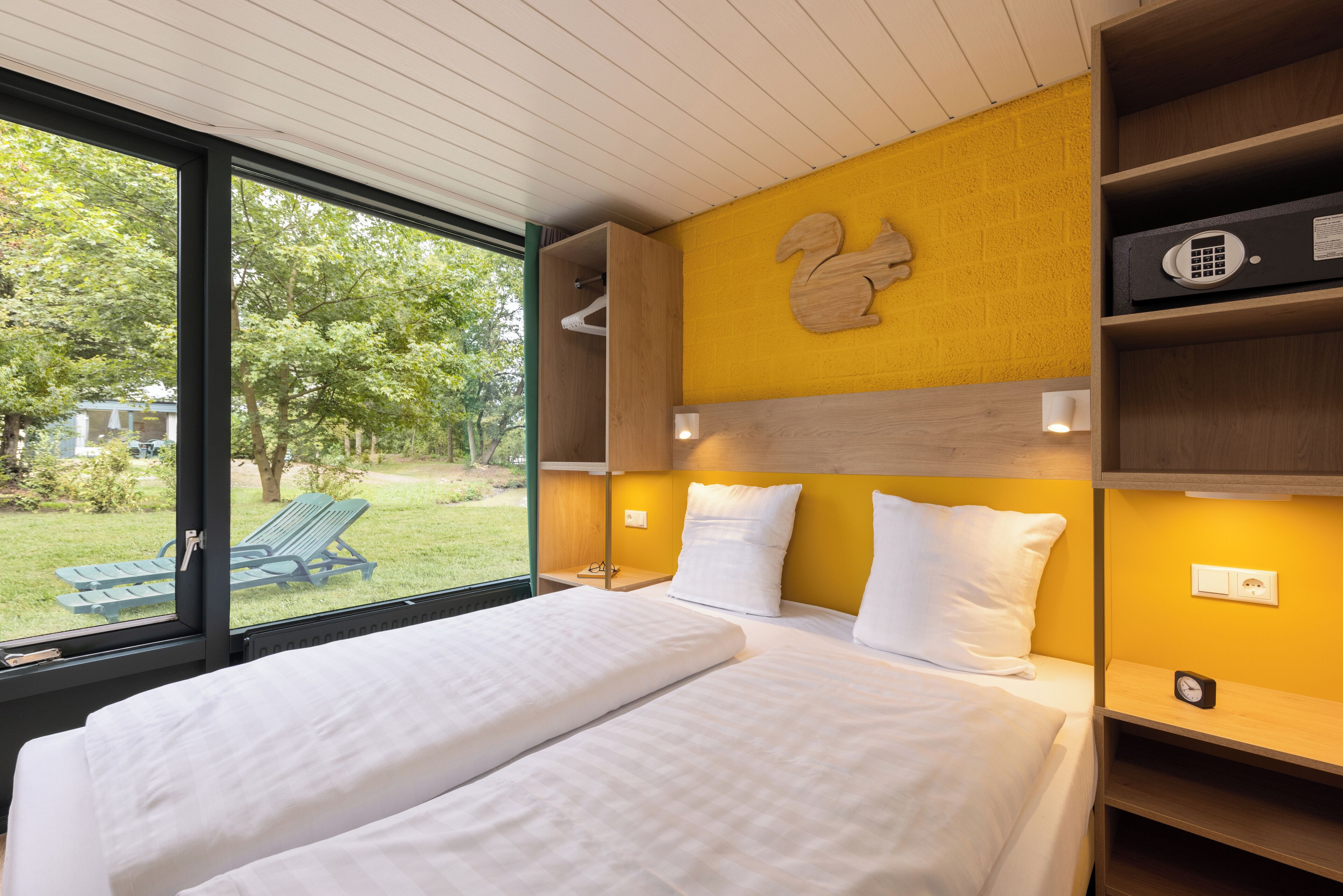 Un dormitorio con una cama grande, cabecera de madera, pared de acento amarilla y vistas a un césped verde y sillones de exterior.