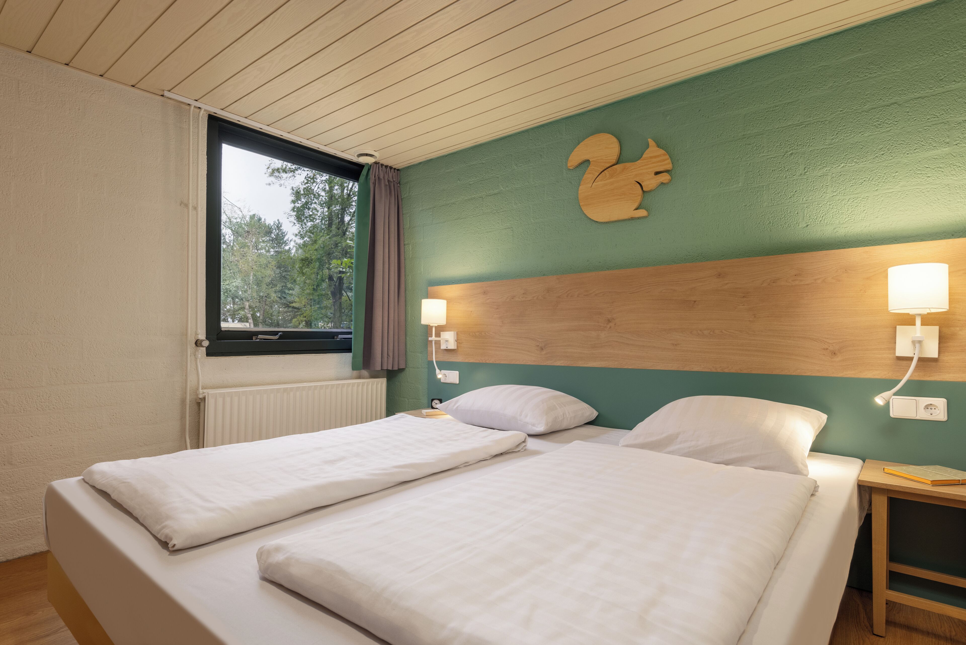 Cama de casal com lençóis brancos, cabeceira de madeira e luminária fixada na parede.