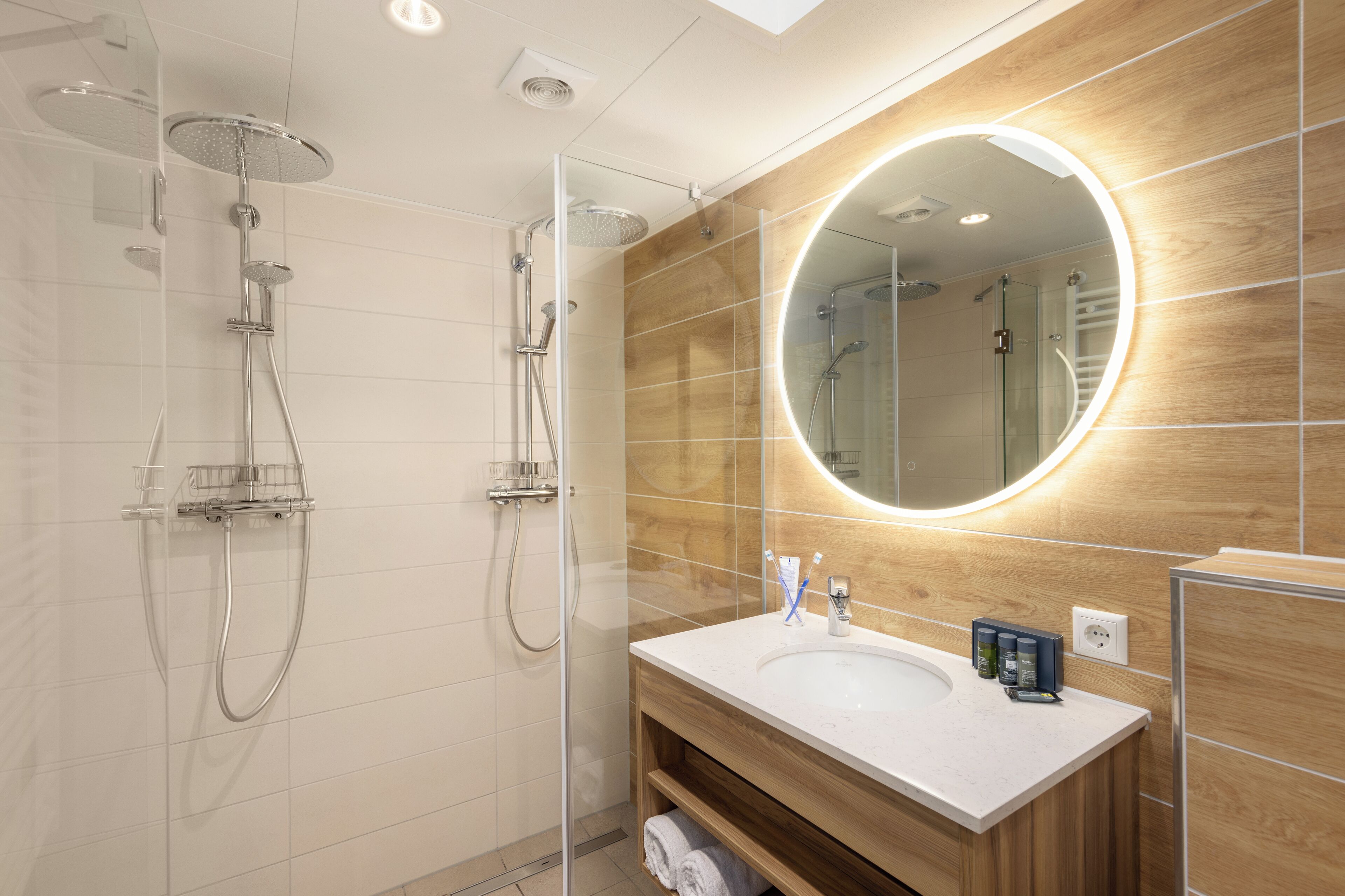 Une salle de bain moderne dotée d’une douche à parois de verre, d’un meuble-lavabo en bois et d’un grand miroir avec un éclairage circulaire.