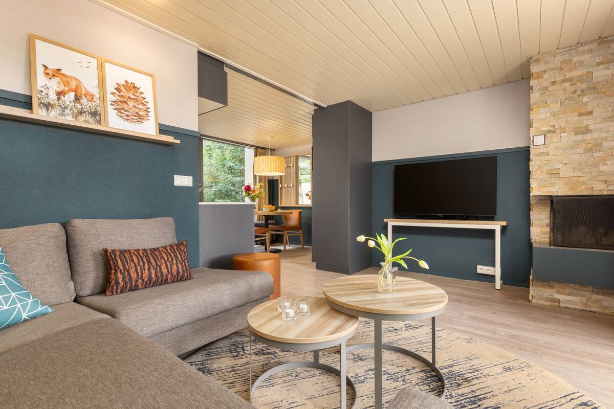A modern living room with a sectional sofa, a flat-screen TV, a stone fireplace, and a dining area in the background.