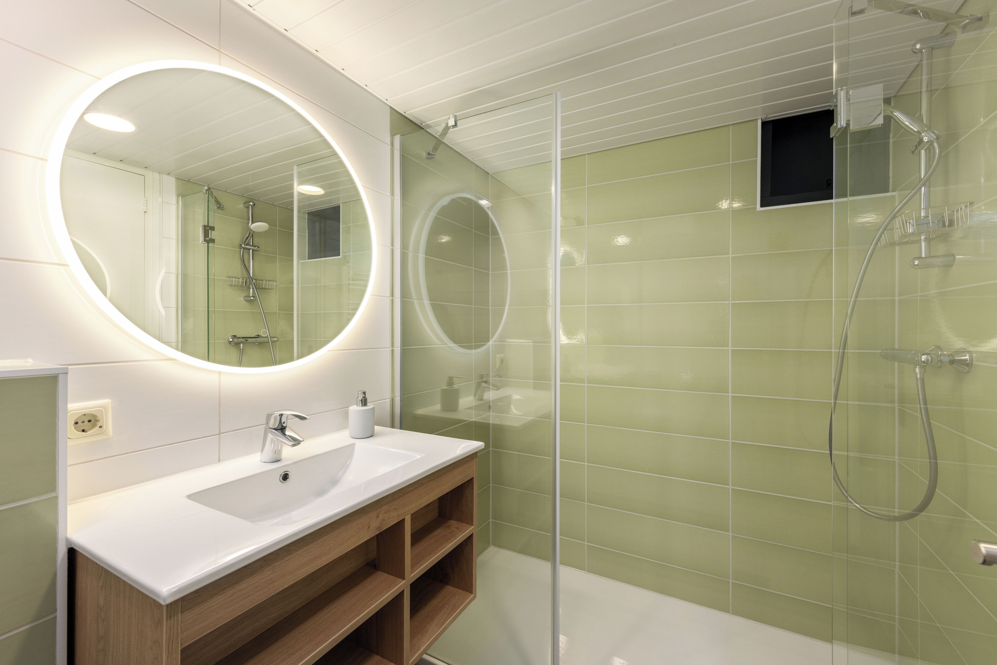 A modern bathroom with a glass-enclosed shower, a white sink with a wooden vanity, and a large mirror with a circular light.