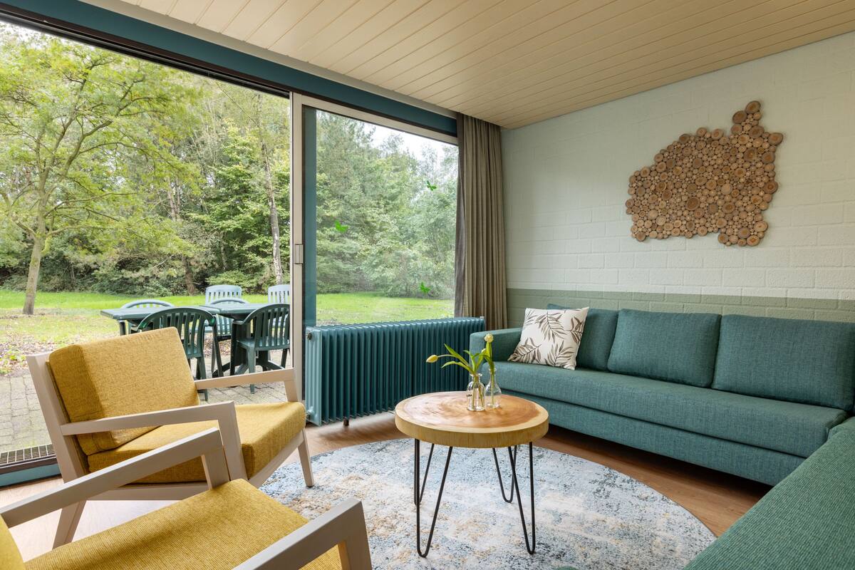 A modern living room with a green sofa, yellow armchair, and a round wooden coffee table. A large window offers a view of a garden and trees.
