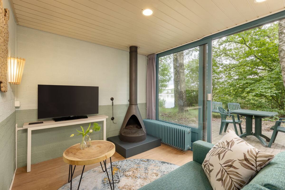 A modern living room with a large TV, a wood-burning stove, a cozy sofa, and a dining area with outdoor seating.