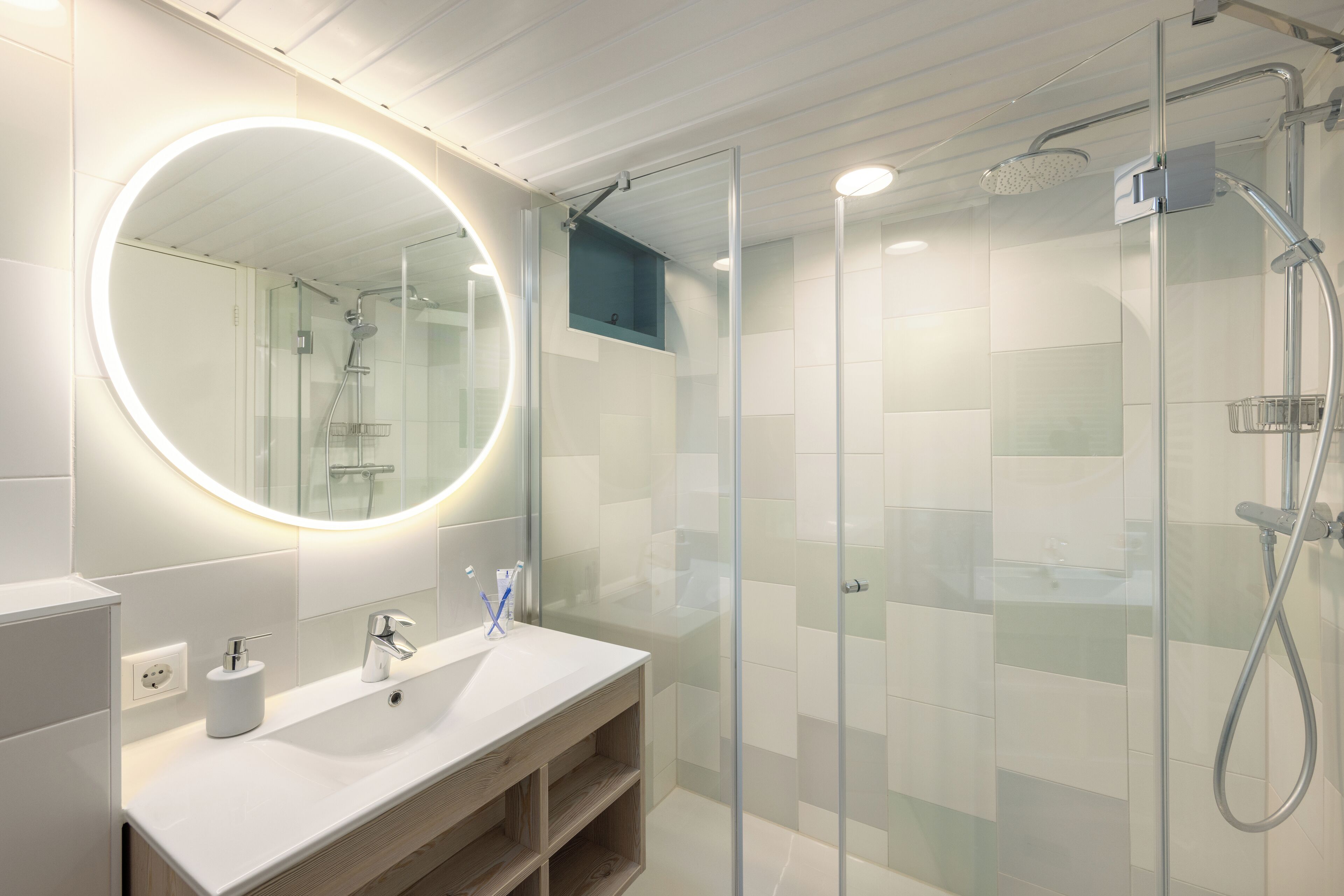 Een moderne badkamer met een douchecabine van glas, een ronde spiegel met verlichting onder de kast, en een witte wastafel met zeepdispenser.
