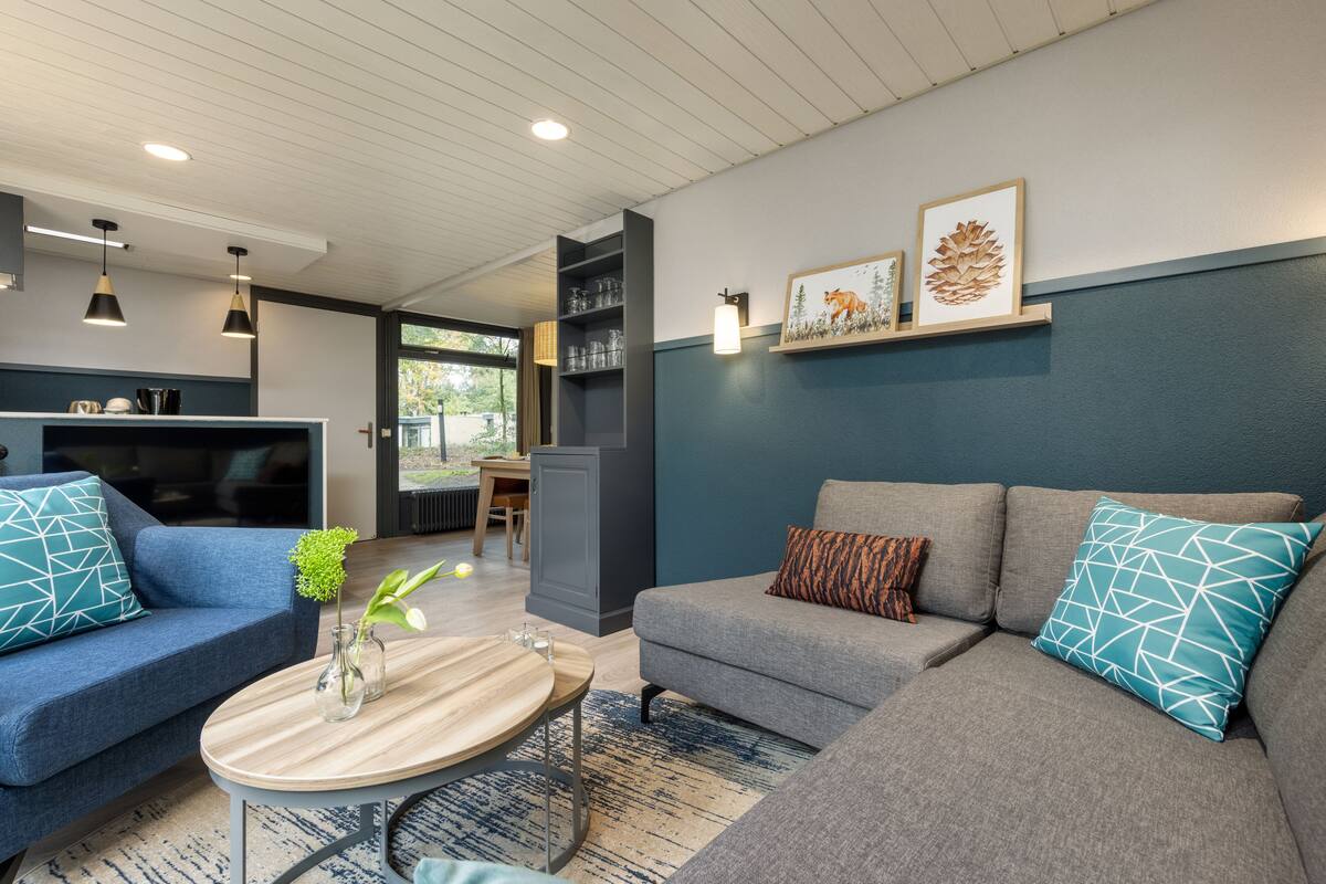 A modern living room with a grey sofa, blue and patterned cushions, a wooden coffee table, and a dining area visible through an open door.