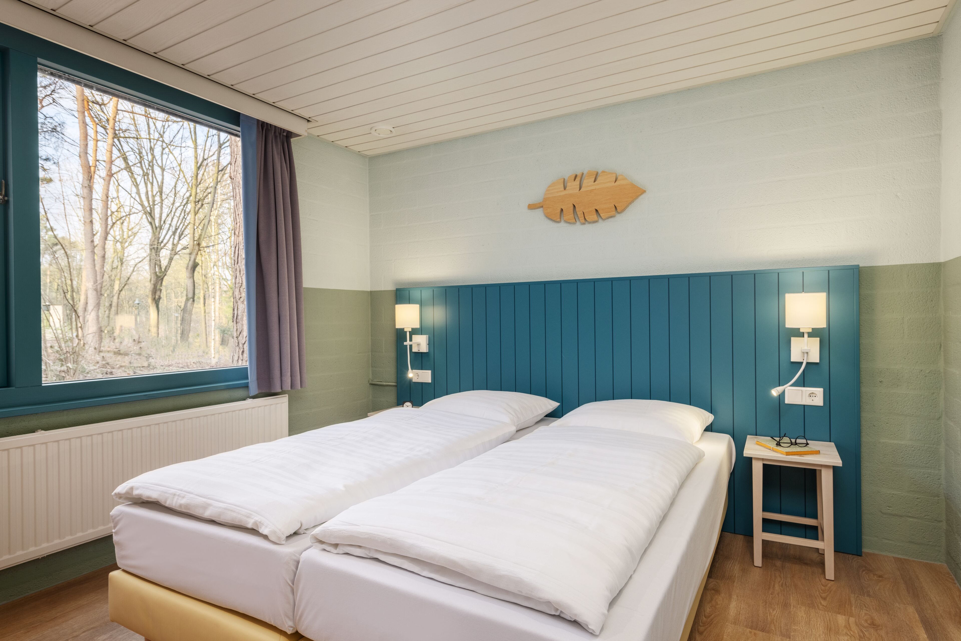 Een tweepersoonsbed met witte beddengoed, een klein nachtkastje met een lamp en een raam met uitzicht op bomen.