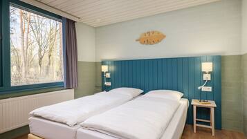Een tweepersoonsbed met witte beddengoed, een klein nachtkastje met een lamp en een raam met uitzicht op bomen.