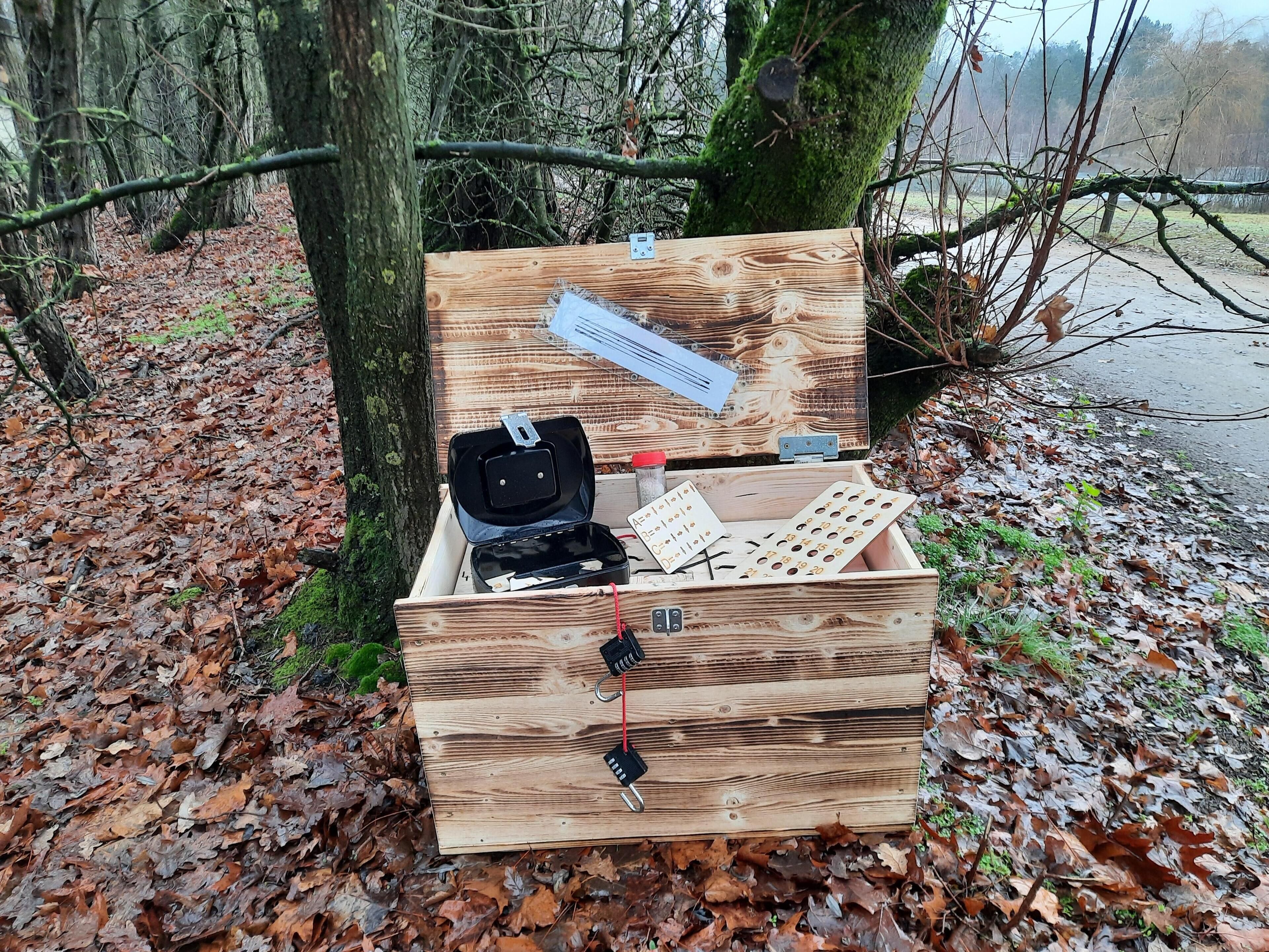 Una scatola di legno con un'etichetta, una borsa nera e una serie di aperture circolari su un terreno boschivo.