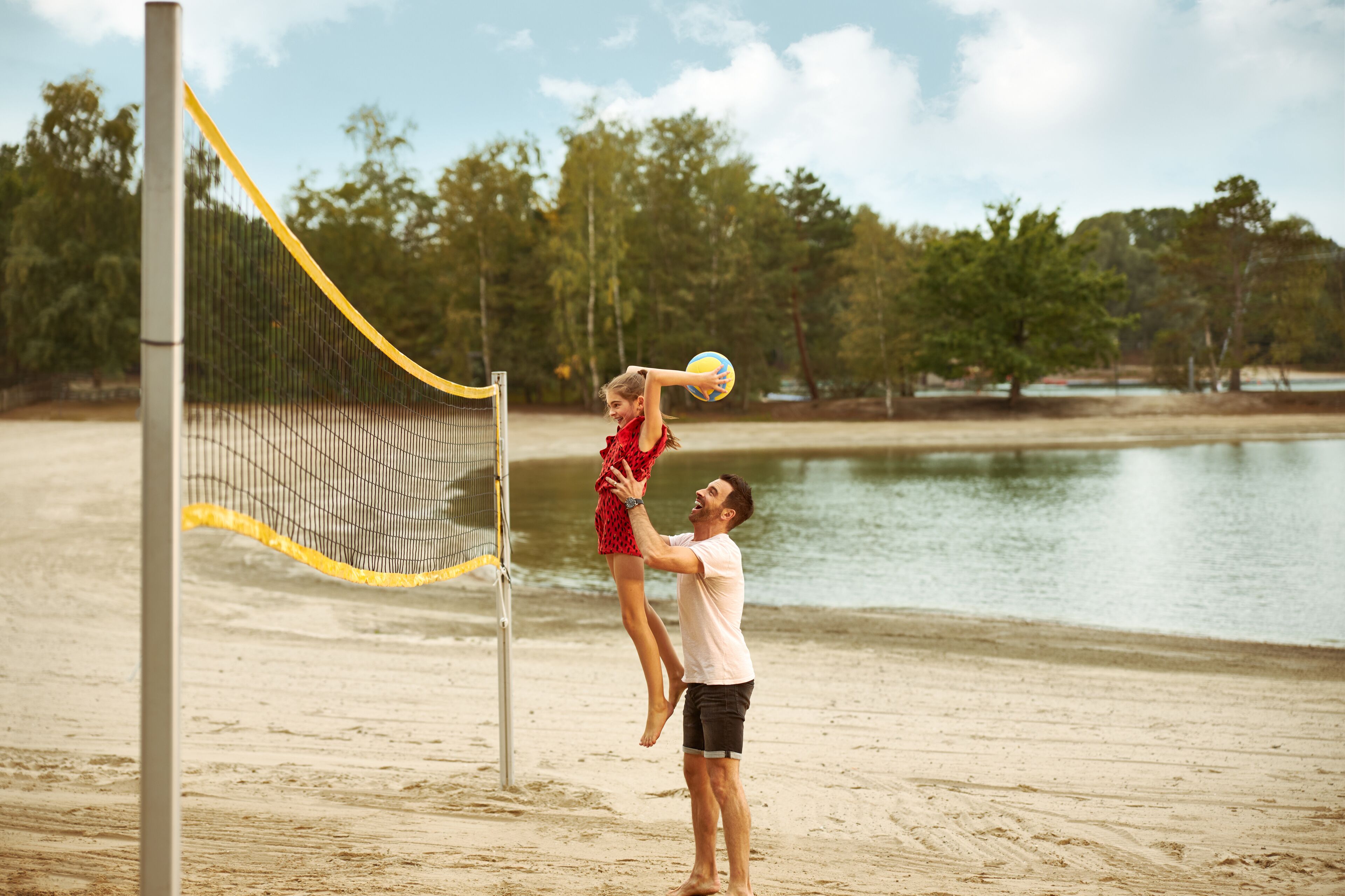 Un match de volley-ball sur une plage de sable, avec un filet, des arbres et une étendue d’eau en arrière-plan.