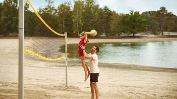 Un match de volley-ball sur une plage de sable, avec un filet, des arbres et une étendue d’eau en arrière-plan.