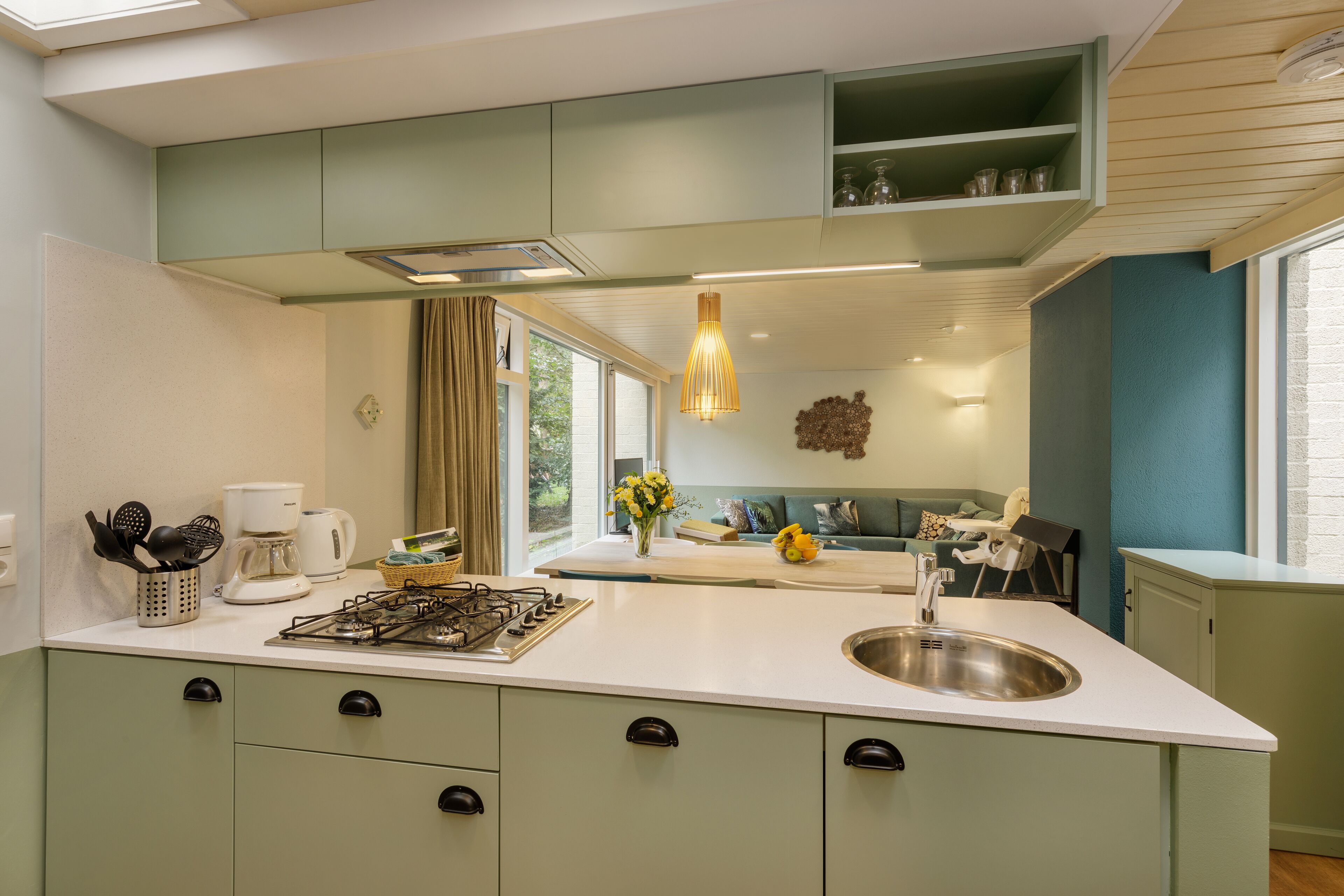 Een moderne keuken met een gasfornuis, spoelbak en groene kastjes. Er is een raam met uitzicht op de omgeving en een hangende lamp.