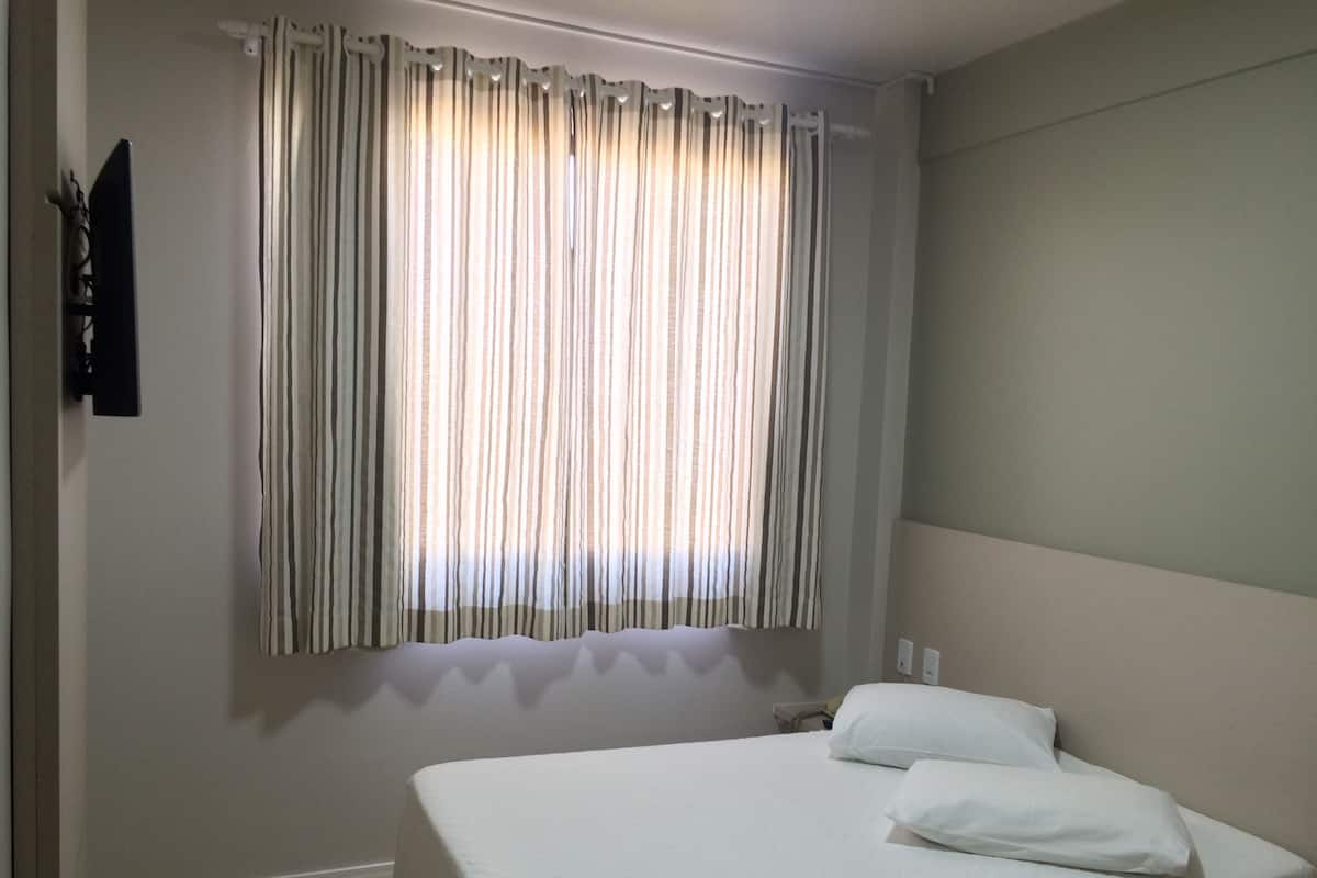 Una cama individual con ropa blanca, una ventana con cortinas a rayas y una pared lisa.
