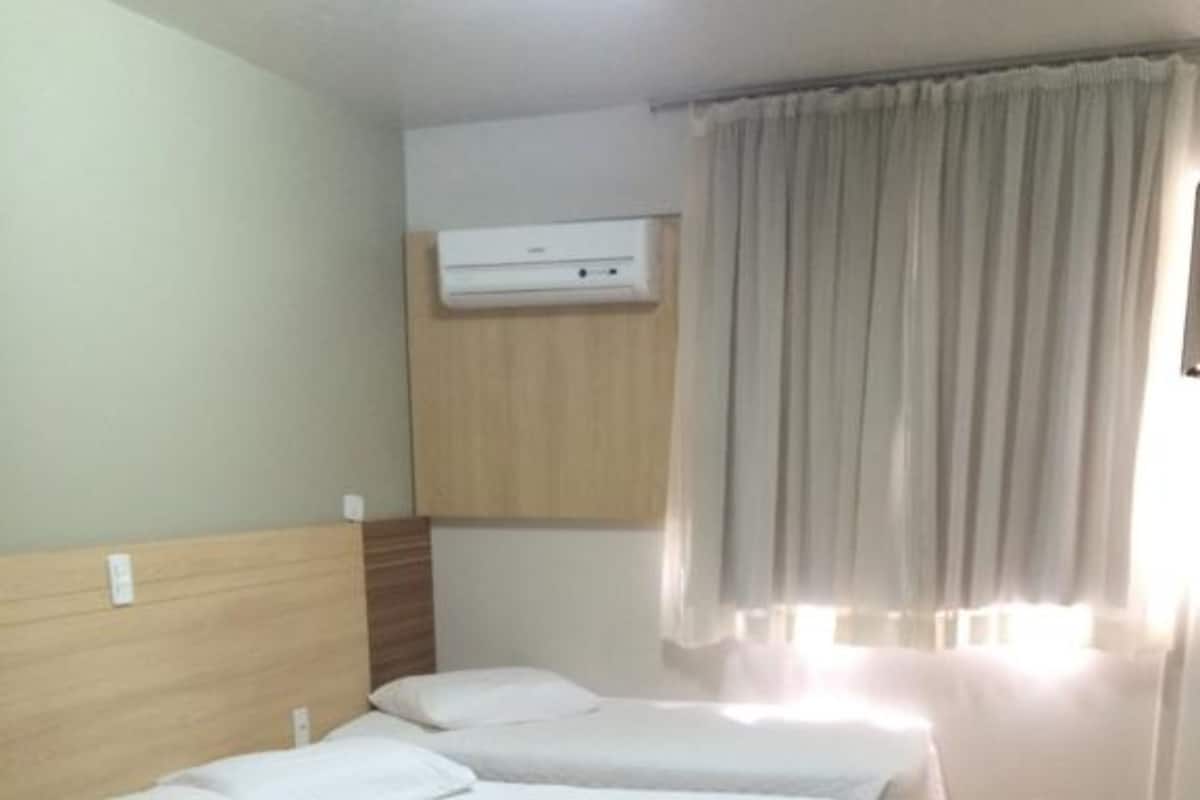 Habitación de hotel con dos camas, cabecero de madera, aire acondicionado instalado en el techo y una ventana con cortinas.