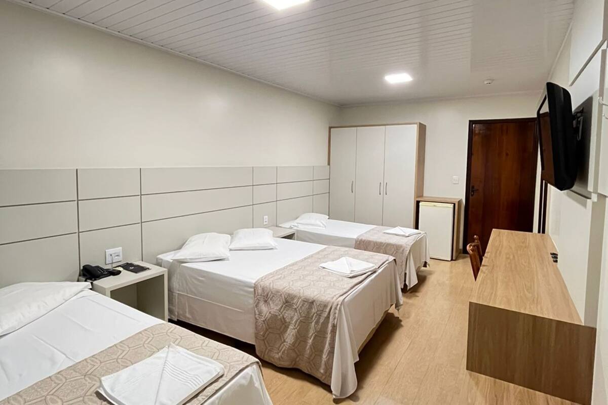 Habitación de hotel con tres camas, un escritorio de madera, un televisor montado en la pared y una puerta que da a otra habitación.