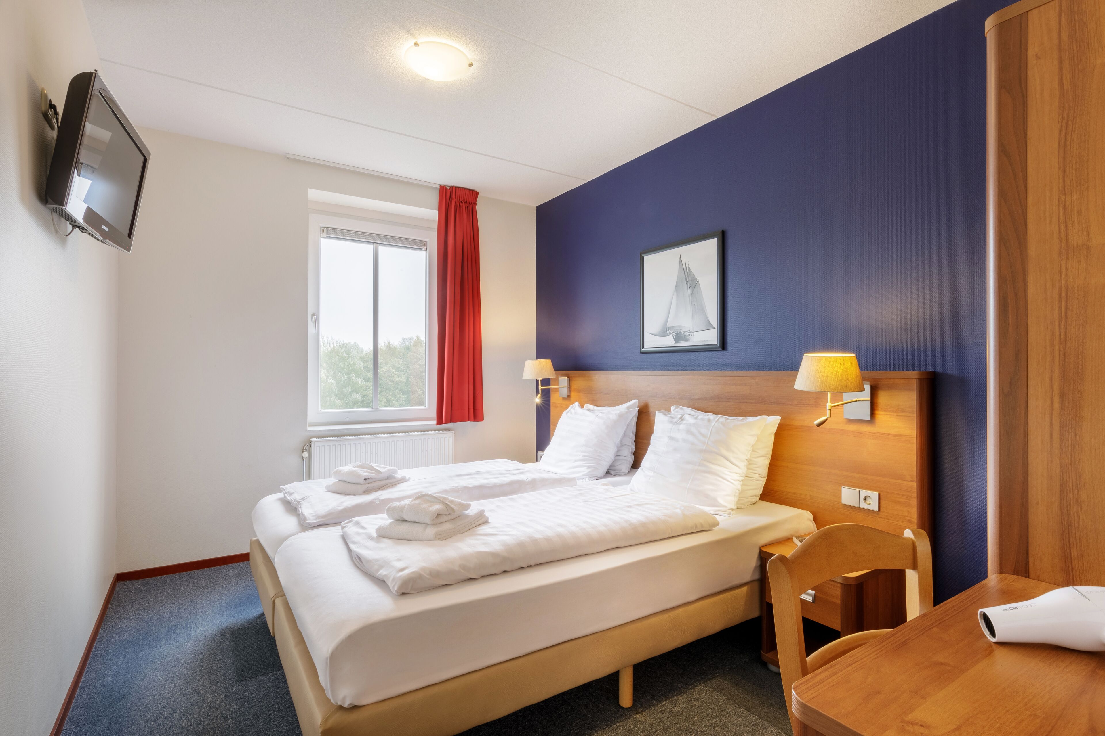 A hotel room with a bed, a desk, a chair, a TV, a window with a red curtain, and a painting on the wall.