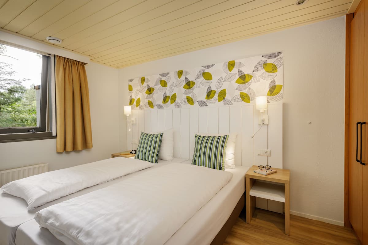A bedroom with two beds, a window with curtains, a wall-mounted light, and a bedside table with a lamp and glasses.
