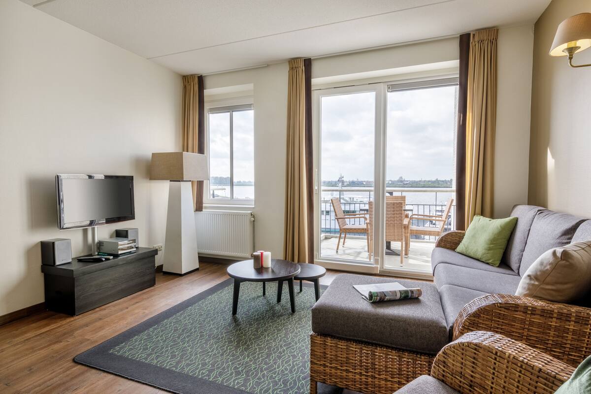 A modern hotel room with a sofa, a TV, a small table, and a view of the city through large windows.