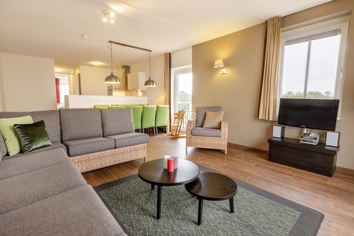 A modern living room with a sofa, armchairs, a coffee table, a TV, and a dining area in the background.