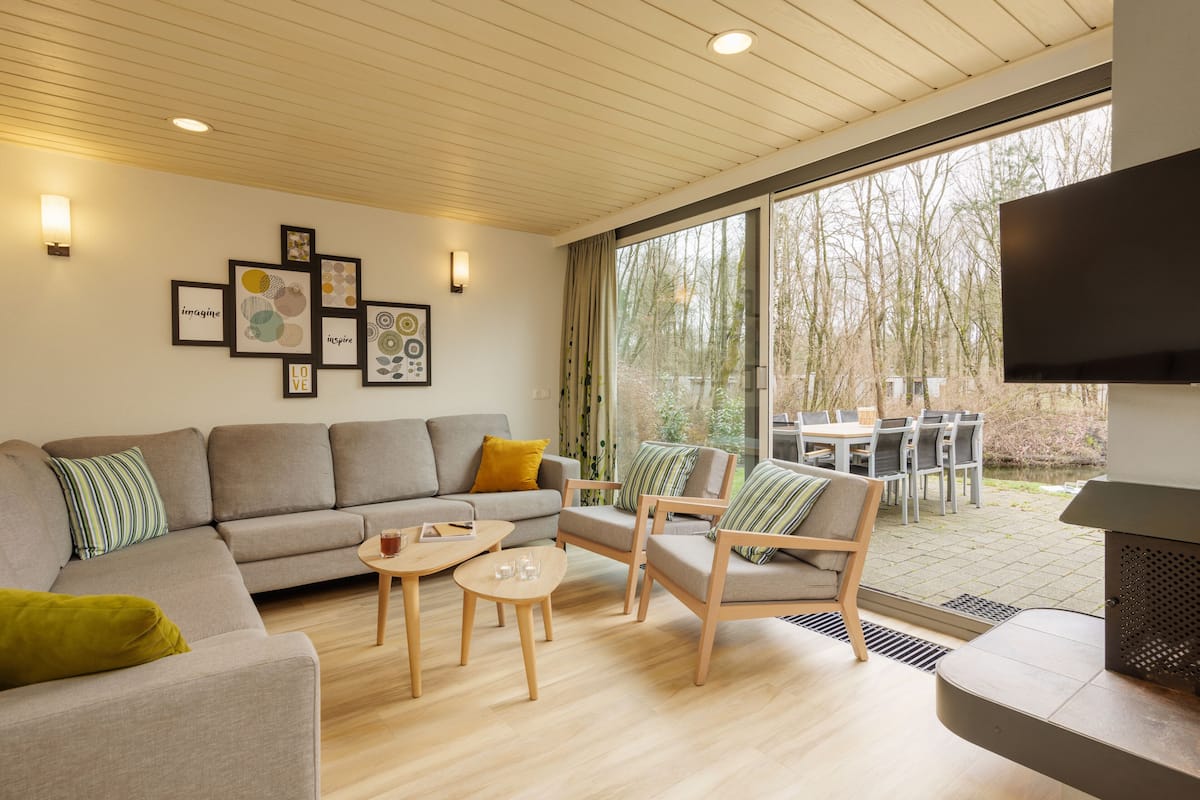 A modern living room with a sofa, armchairs, a coffee table, and a TV. There are framed pictures on the wall and a window with a view of trees.